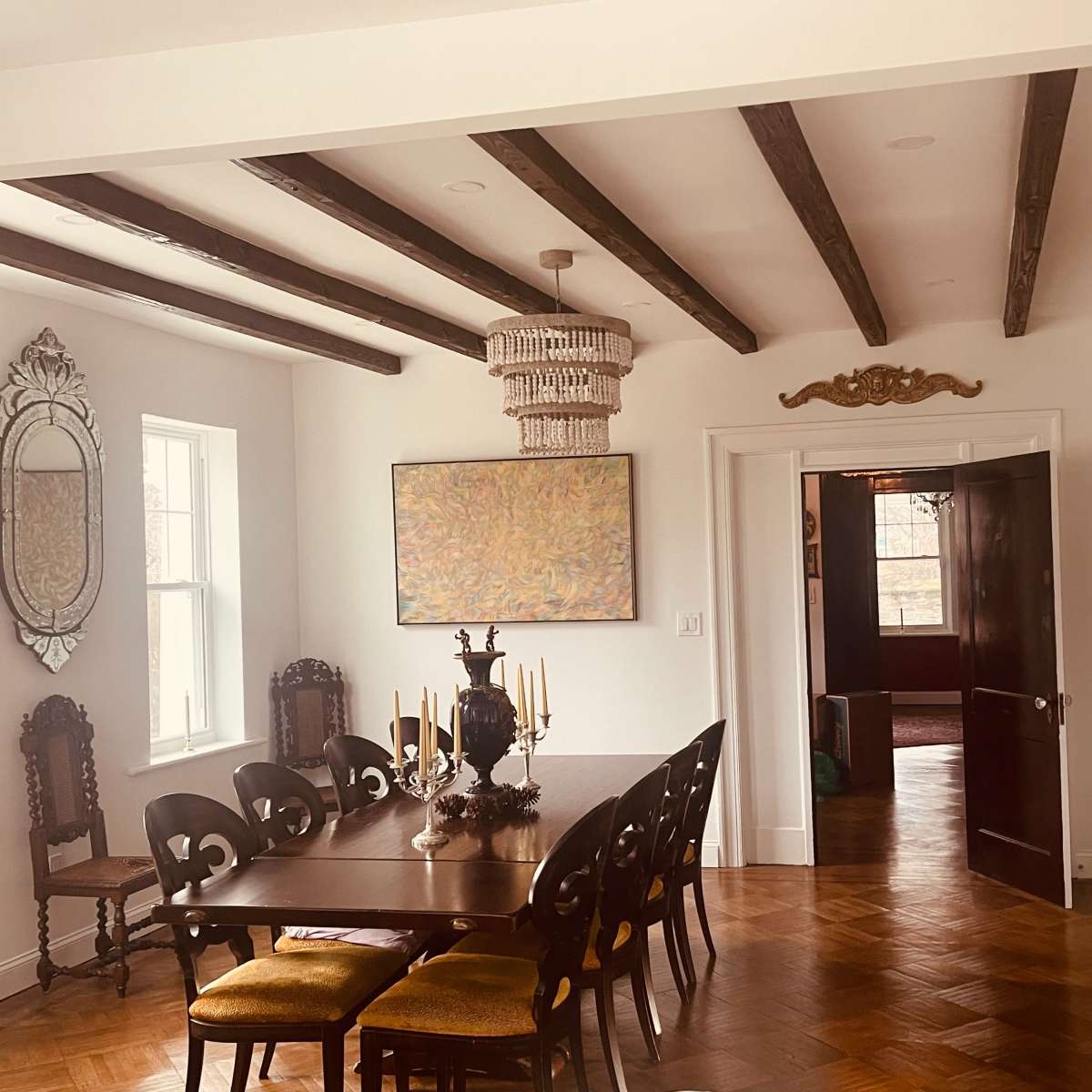 A dining room features a long wooden table surrounded by chairs, with a chandelier above and an ornate mirror on the wall.
