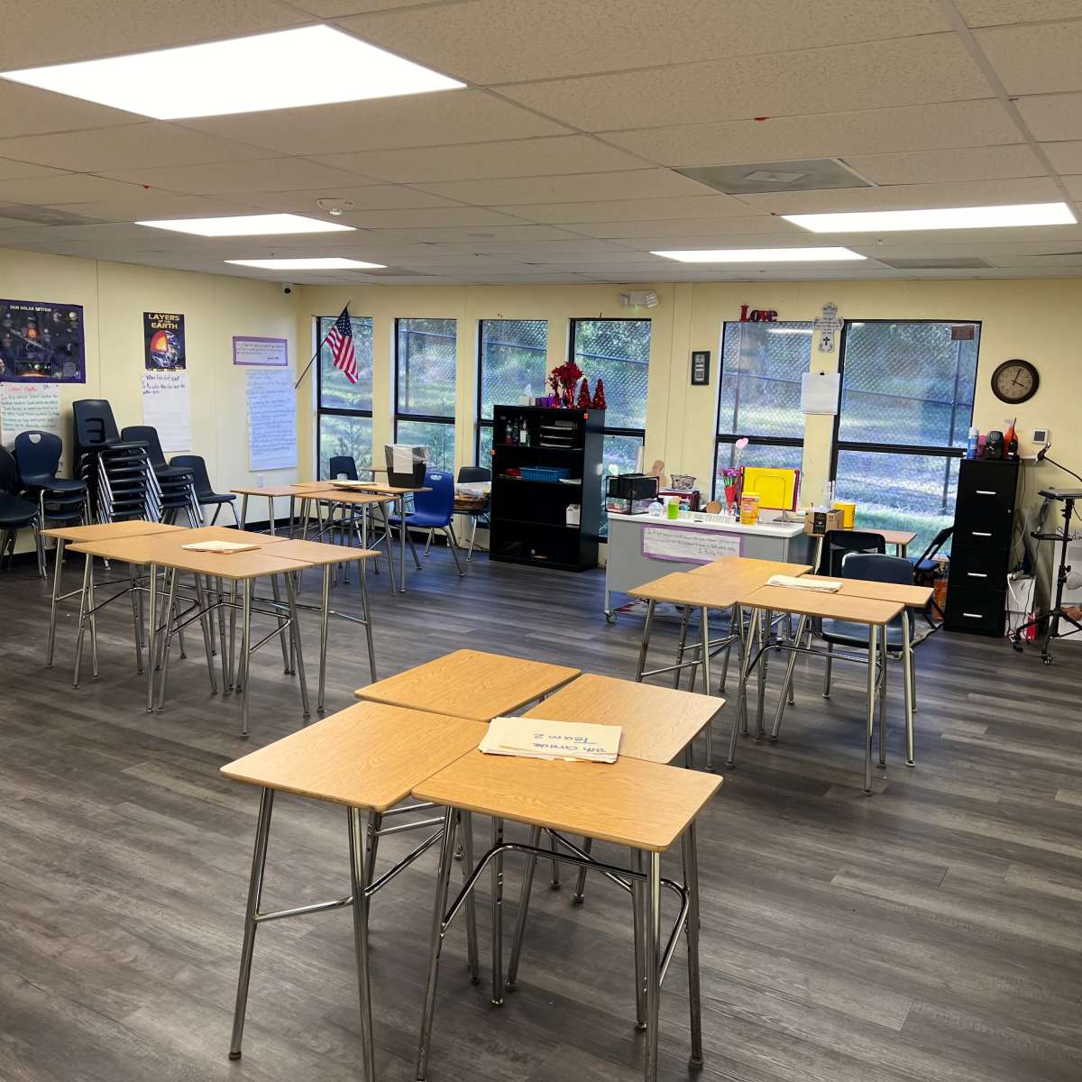 A classroom with several empty tables arranged in rows and various educational resources displayed on shelves and walls.