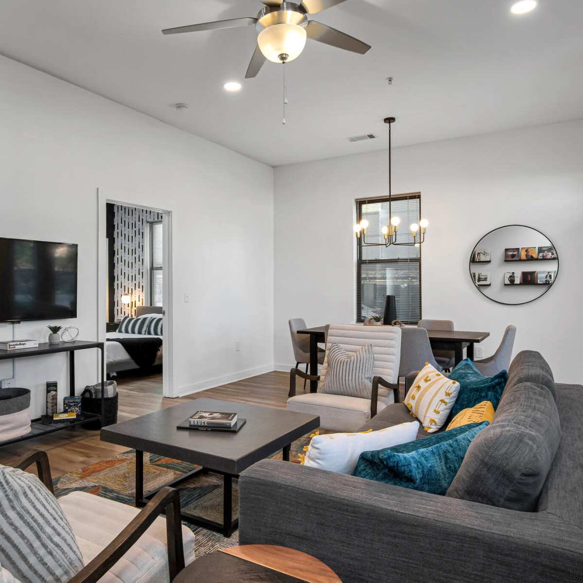 A modern living room with a sectional sofa, a coffee table, a television on a stand, and a dining area with a round table and chairs in the background.