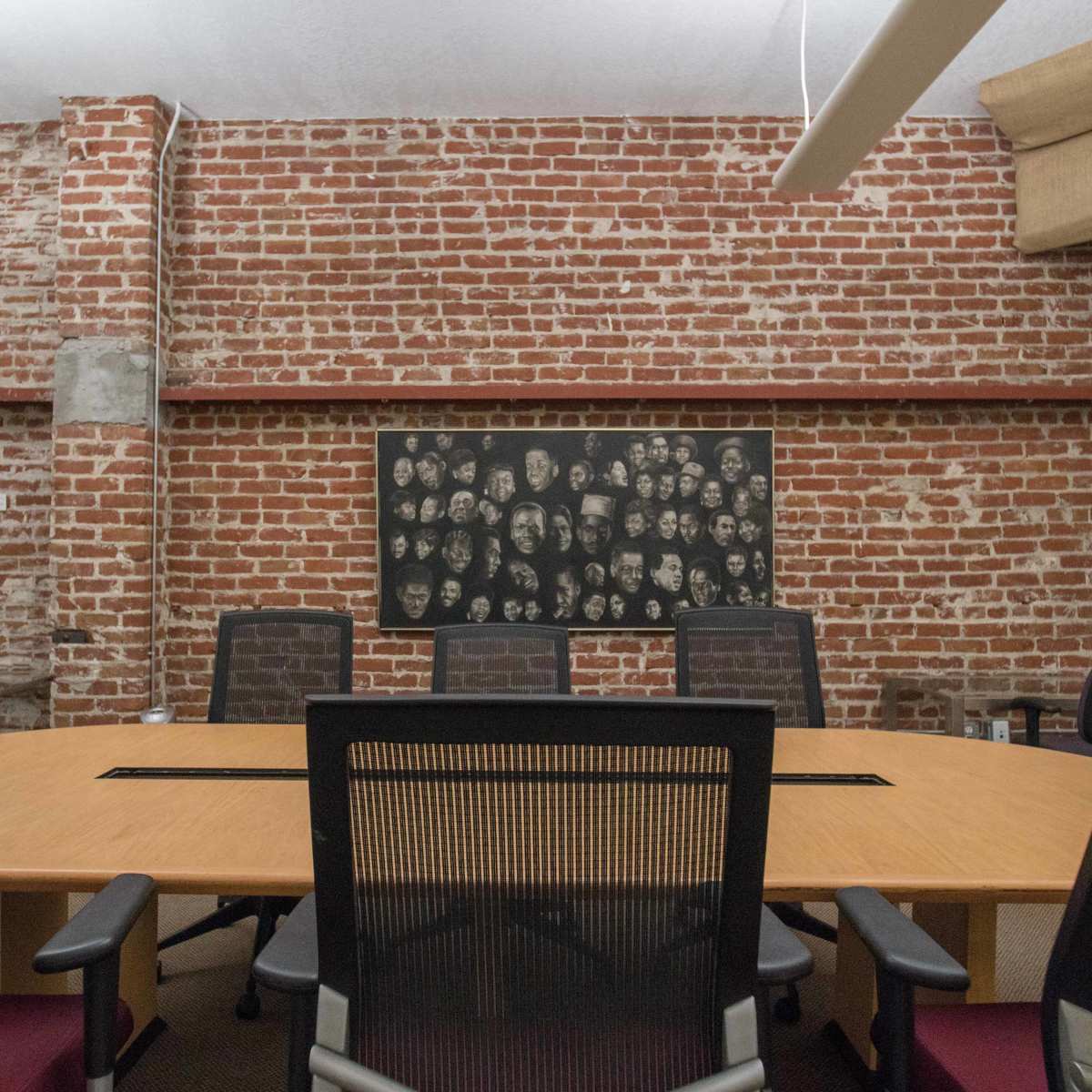 A conference room features a large wooden table surrounded by black chairs, with a red brick wall decorated with framed art and a collage of faces.