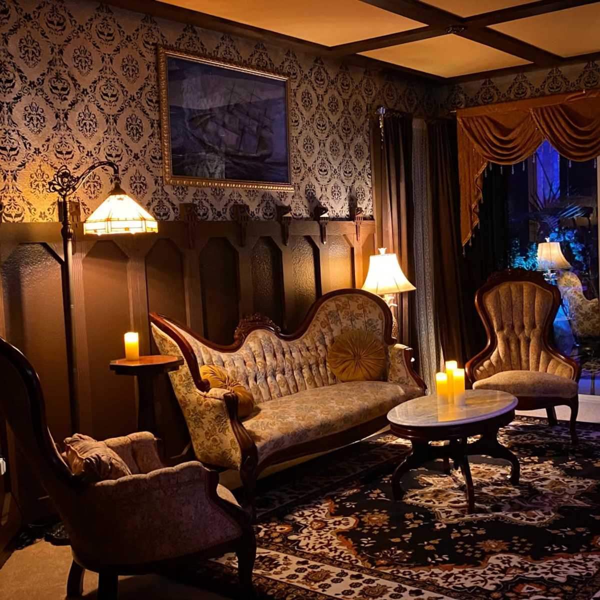A dimly lit parlor with vintage furniture, including a patterned sofa and two armchairs, illuminated by table lamps and candles, against a backdrop of ornate wallpaper and a decorative rug.