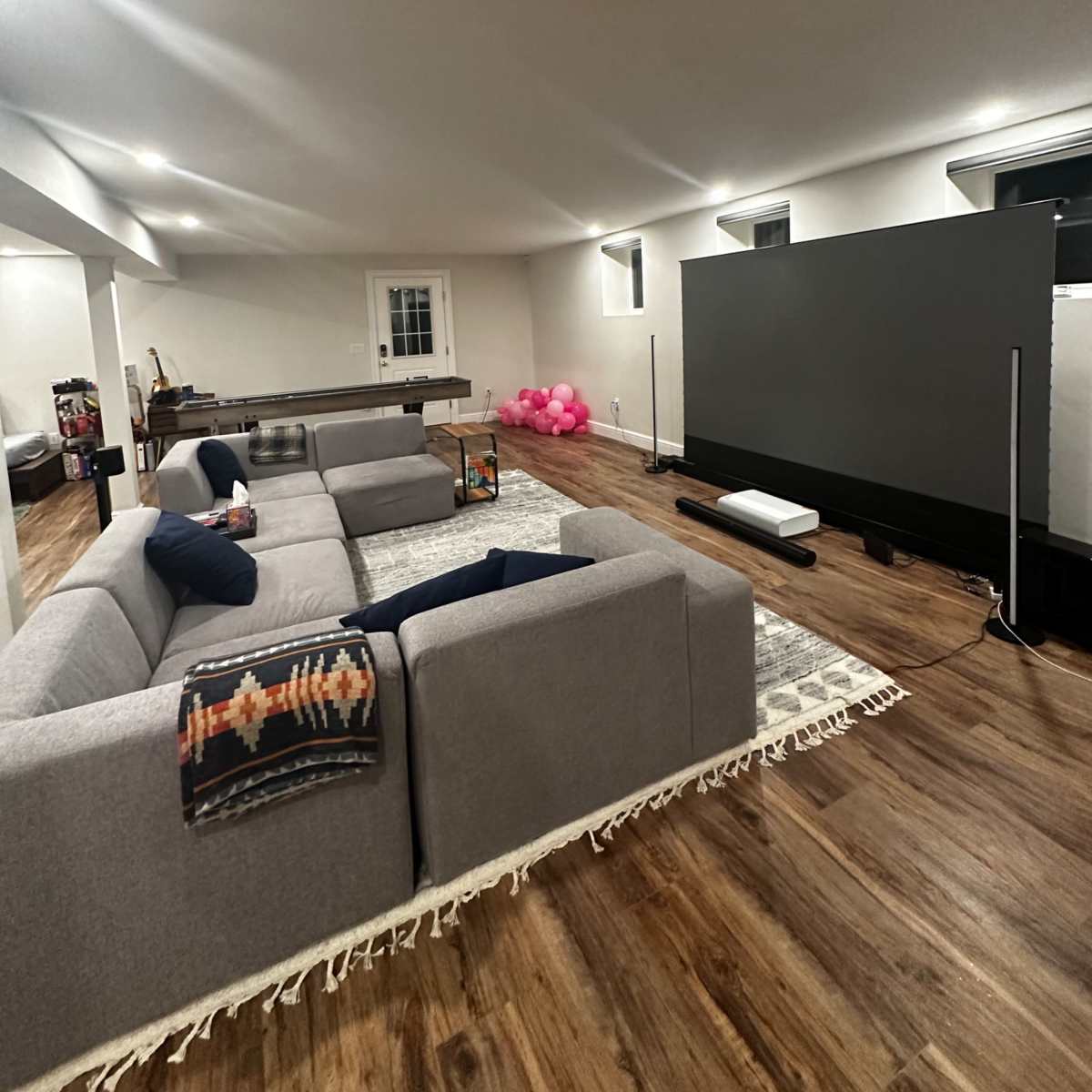 A spacious, well-lit basement living area featuring a large gray sectional sofa, a wooden coffee table, and a screen set up against one wall, with a cluster of pink balloons in the corner.