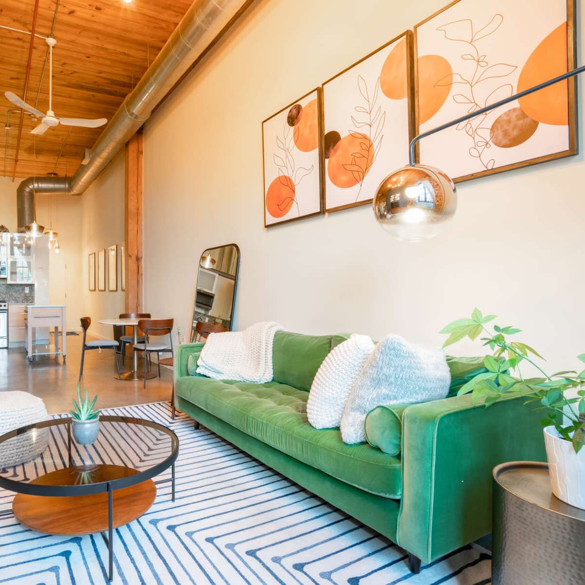 A modern living room features a green sofa, a round coffee table, and artwork displayed on the wall, with visible wood beams and appliances in the background.