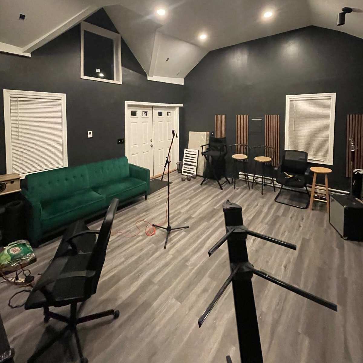 A dimly lit rehearsal space with a green couch, a microphone stand, several stools, and various musical equipment along the walls.