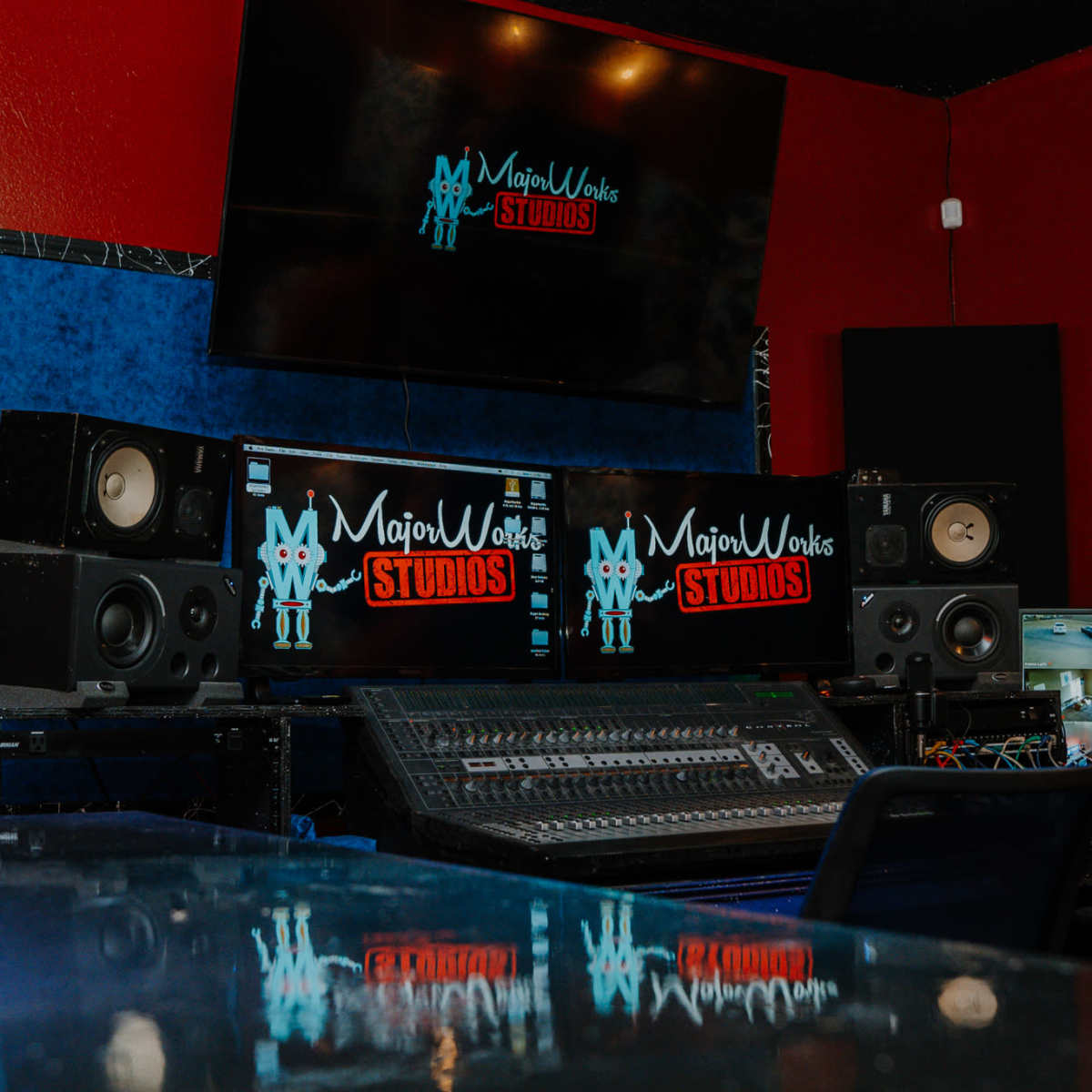 A music recording studio setup with large speakers, a mixing console, and monitors displaying the "MajorWoks Studios" logo on red and blue walls.