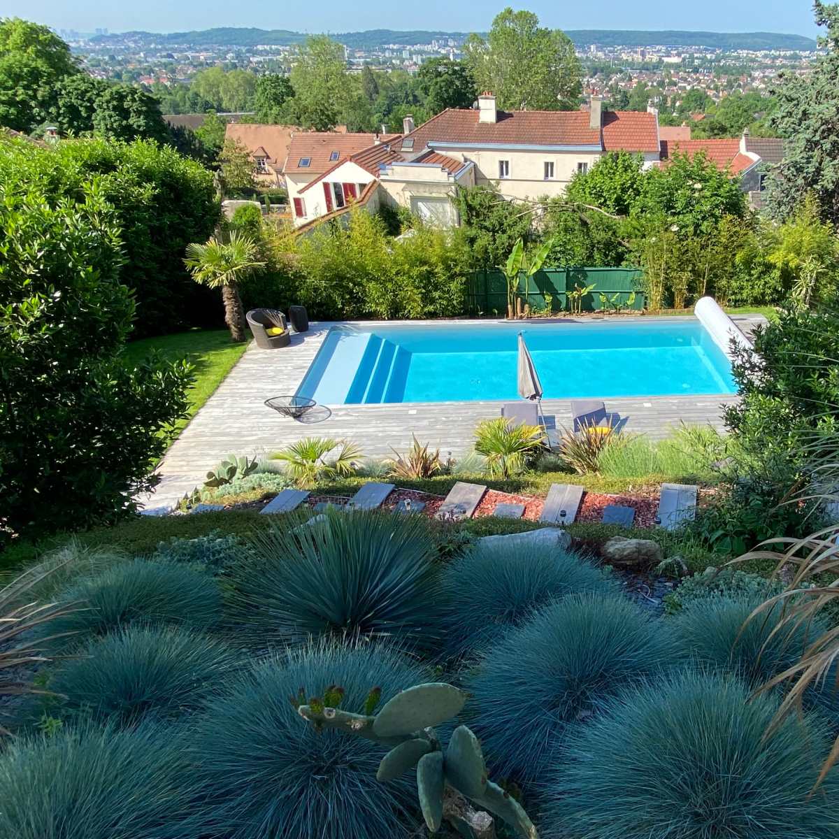 A clear blue swimming pool surrounded by a landscaped garden with various plants and trees, set against a backdrop of distant hills and houses.