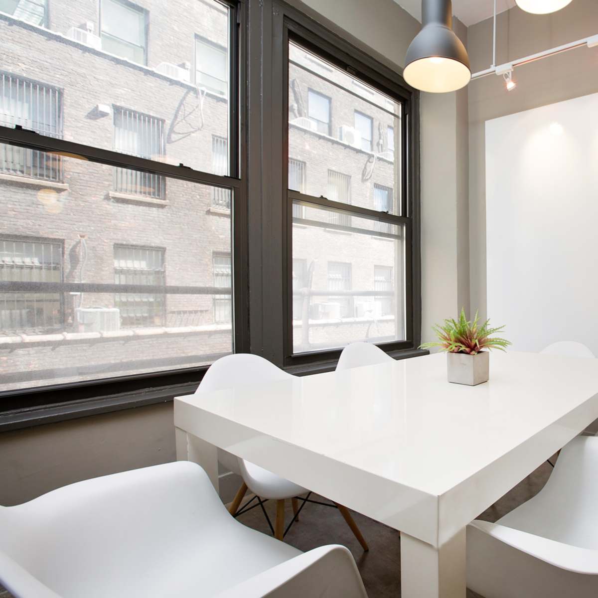 A modern conference room with a white table surrounded by four white chairs, illuminated by overhead lights and featuring large windows.