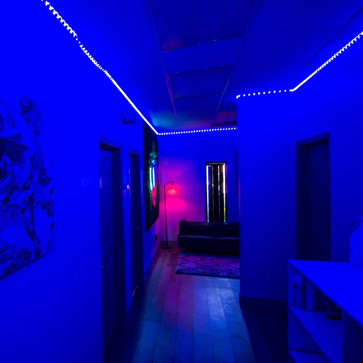 A dimly lit hallway features blue LED lighting, an abstract wall painting, and a red lamp illuminating a couch at the end.