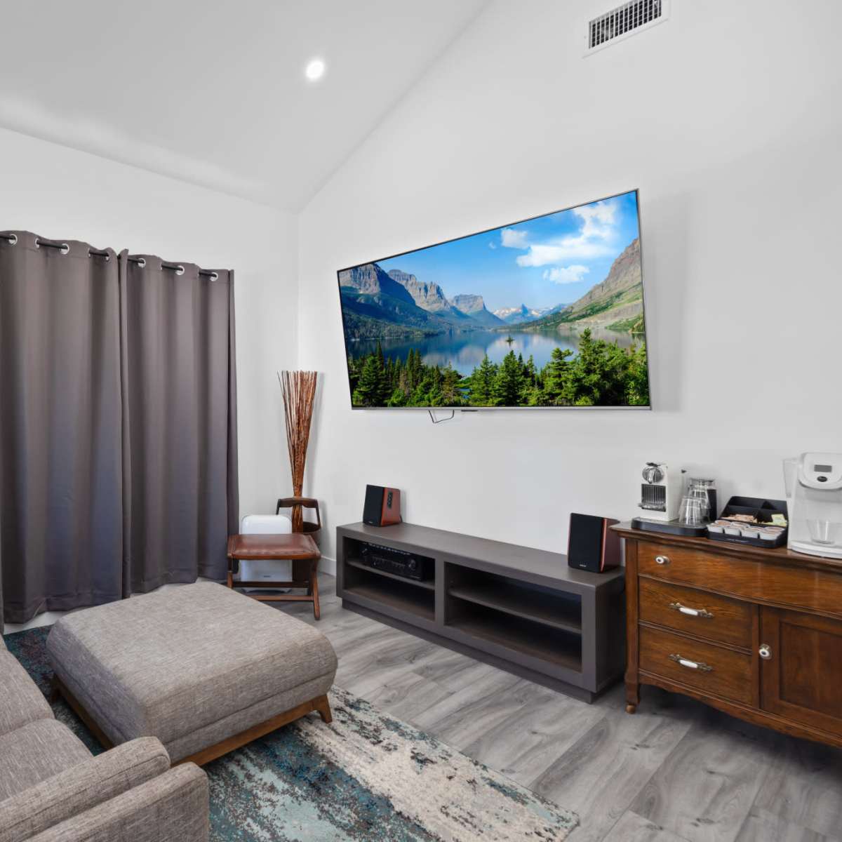 The living room features a gray sectional sofa, a large wall-mounted television displaying a scenic landscape, and dark curtains beside a wooden cabinet.
