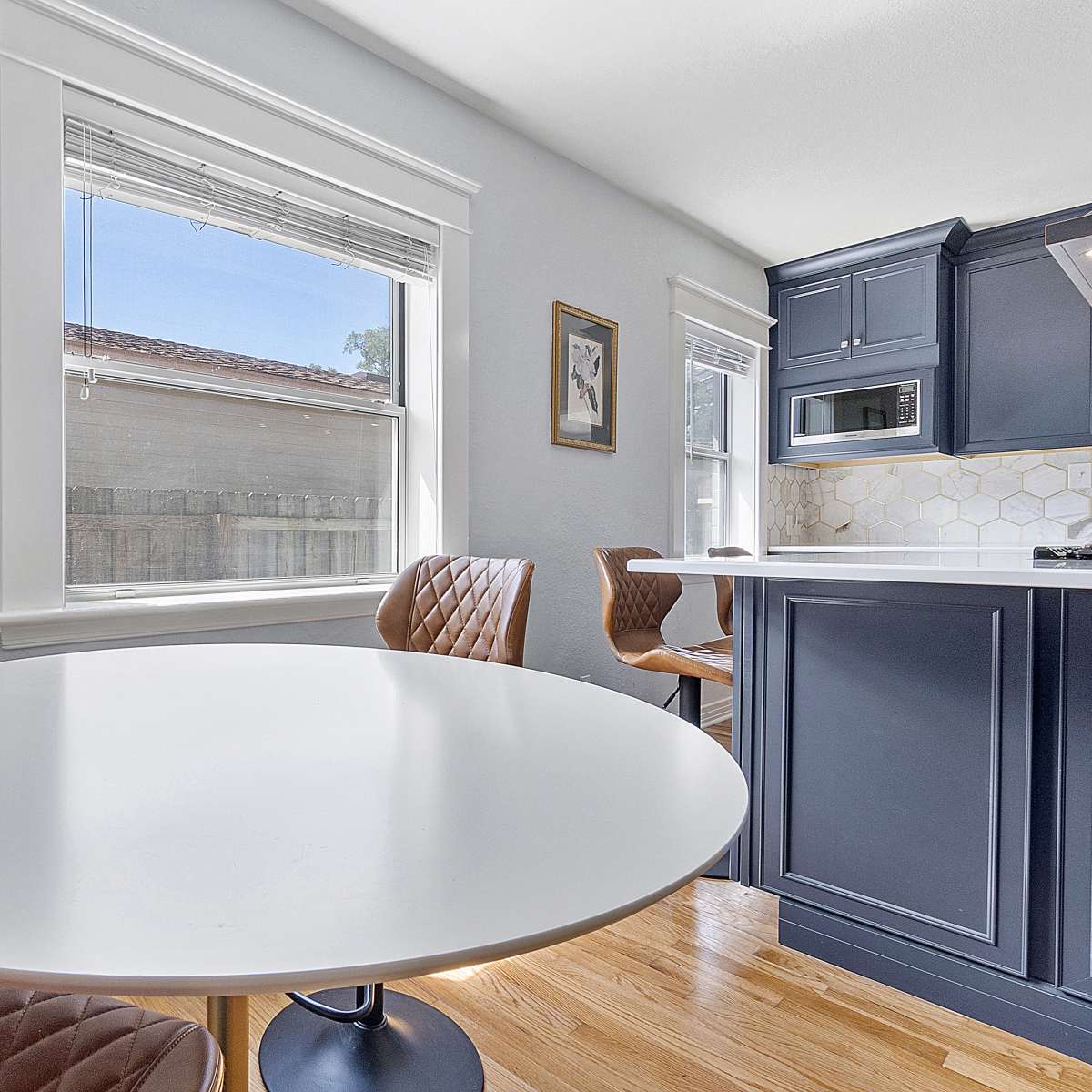 A round white dining table is positioned near a window in a kitchen with navy blue cabinets.