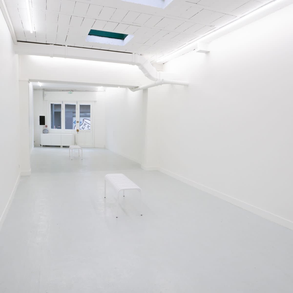 A minimalist gallery space with white walls, a light gray floor, and a few transparent benches.