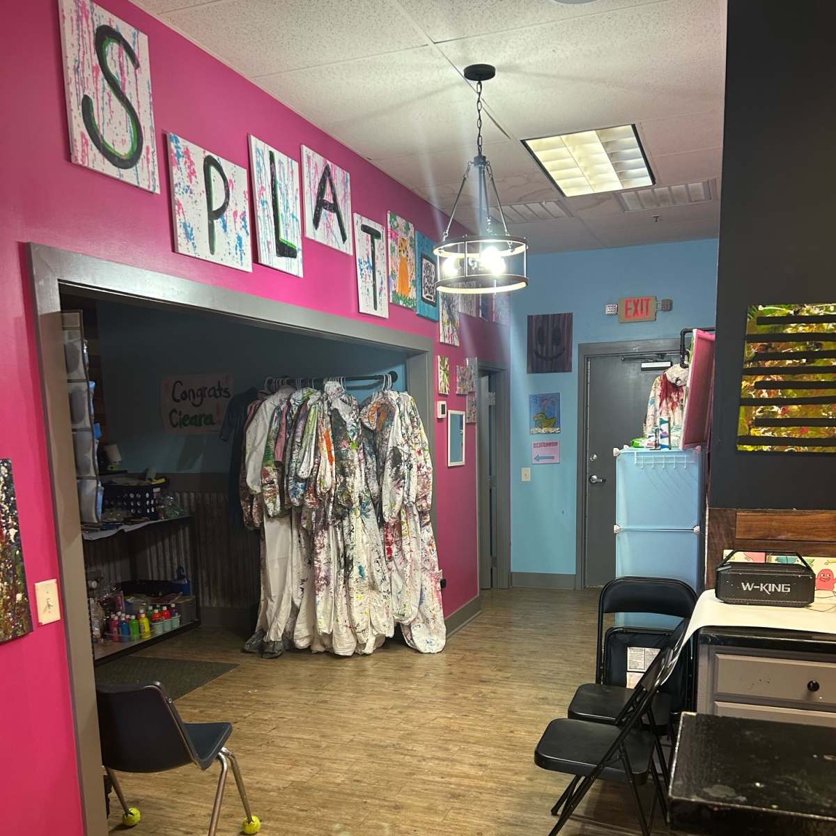 A brightly colored art studio with a corridor leading to a room adorned with splattered paint and hanging artist smocks.