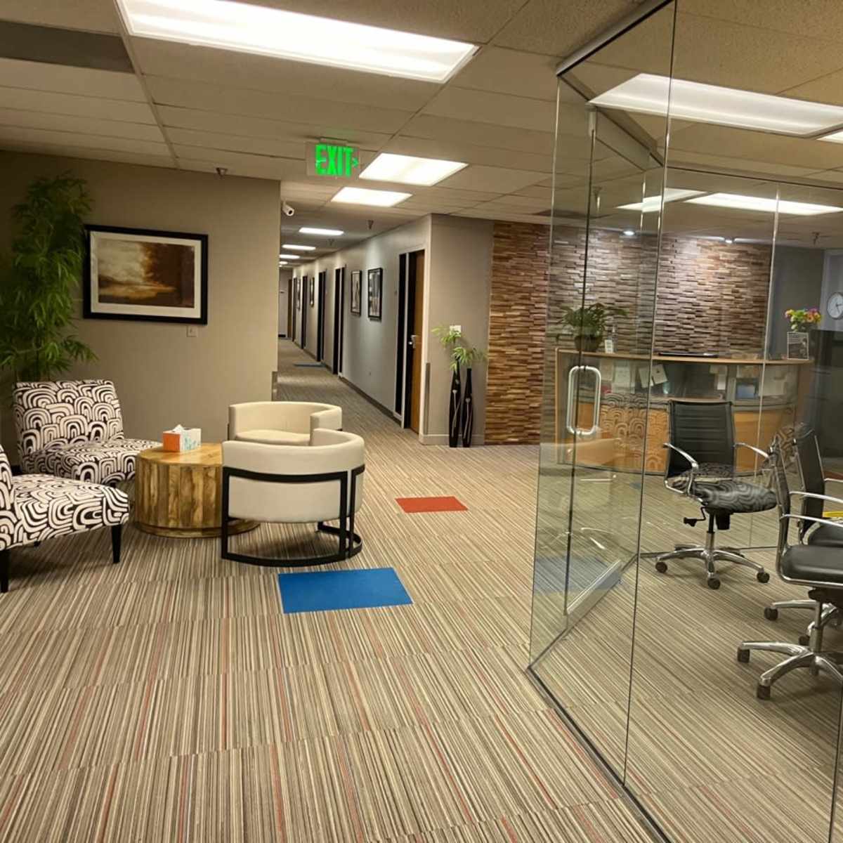 A modern office space featuring glass walls, a conference table, chairs, and a waiting area with patterned and solid color seating.
