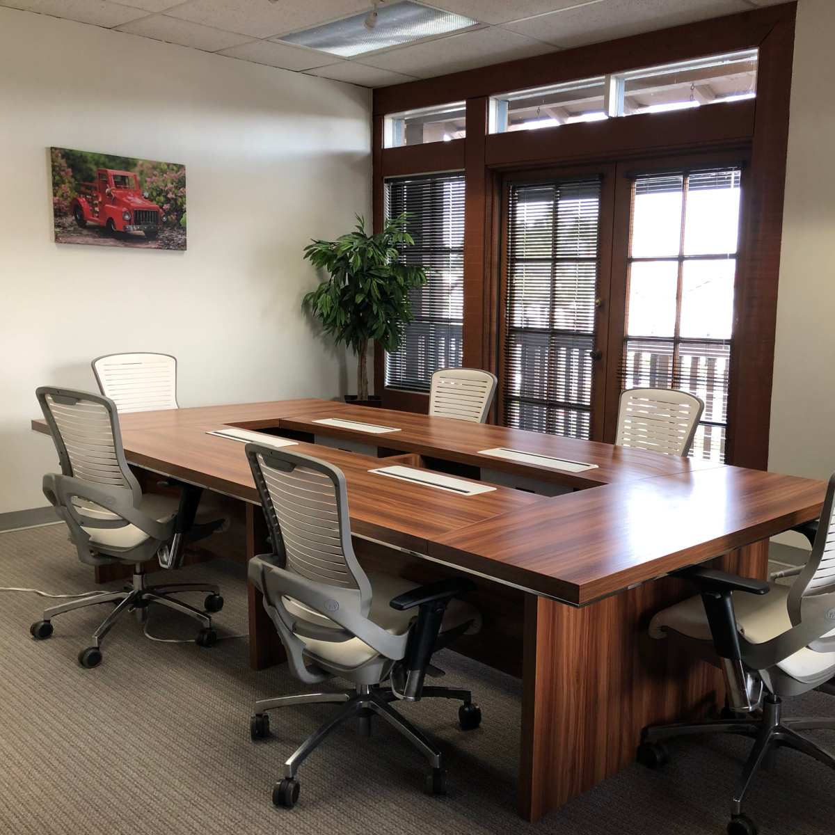 A conference room with a large wooden table surrounded by five ergonomic chairs, and large windows with blinds letting in natural light.