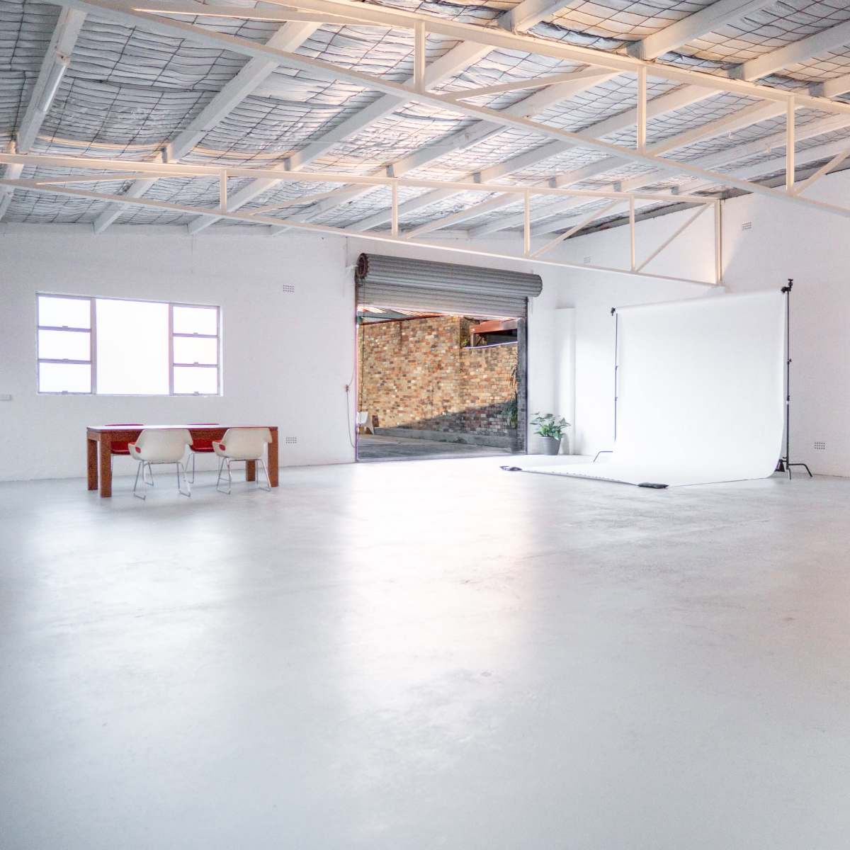 A spacious, empty industrial-style room with a white backdrop set up, a table, and two chairs.