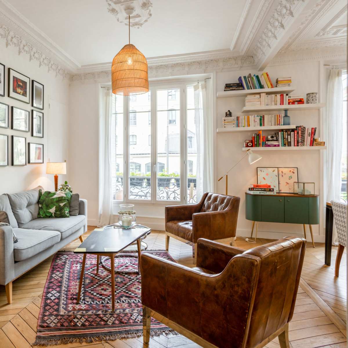 A cozy living room features a gray sofa, wooden armchairs, a coffee table, and bookshelves, with large windows allowing natural light.