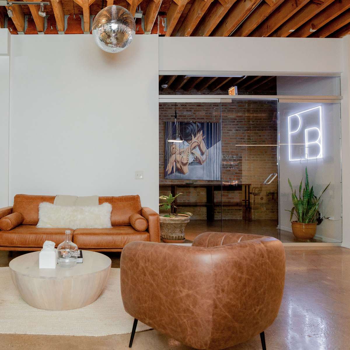 A modern living space with a leather sofa, two brown armchairs, a circular coffee table, and a plant, set against a backdrop of exposed wooden beams and a neon sign.