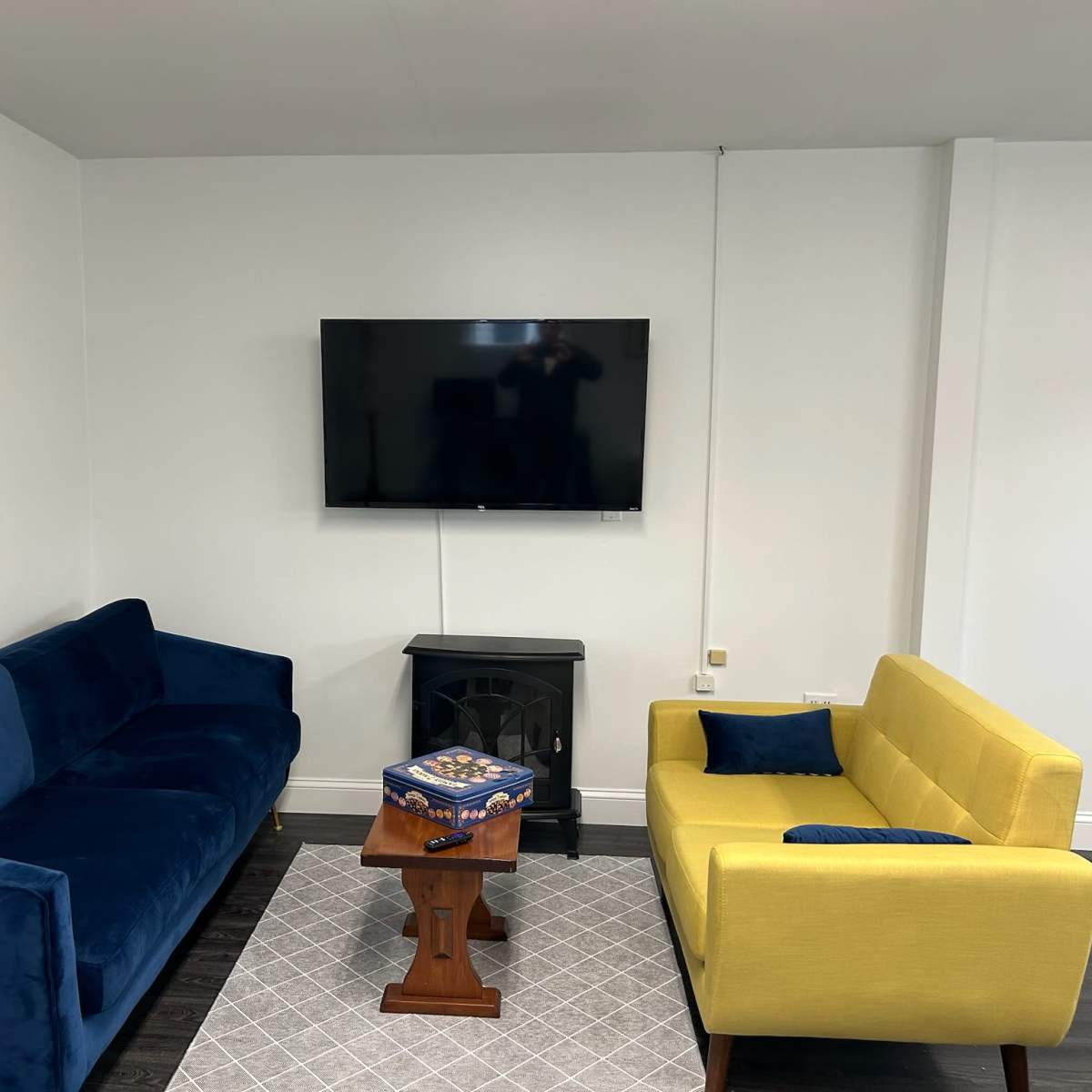 A living room with a blue sofa and a yellow sofa facing a wall-mounted television, along with a small wooden table and a decorative rug on the floor.