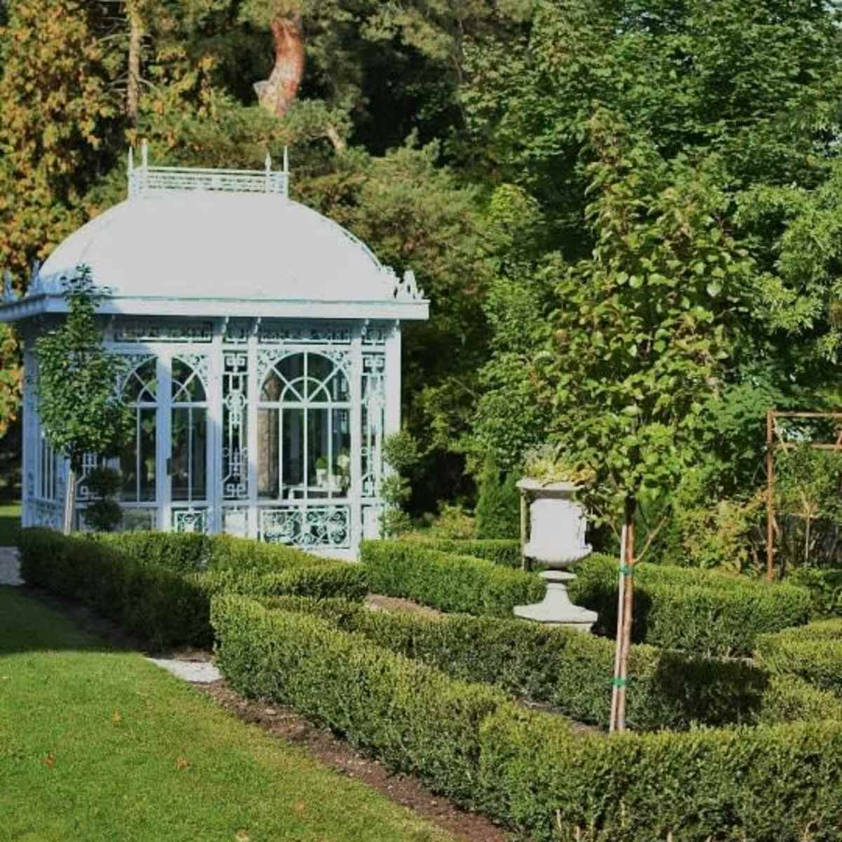 A decorative glass gazebo surrounded by neatly trimmed hedges and trees in a lush garden setting.