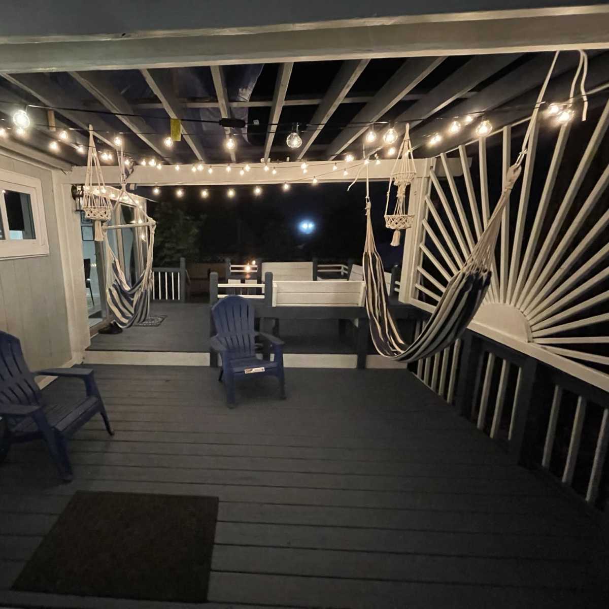 A nighttime view of a porch with hanging lights, two hammocks, and chairs arranged on a wooden floor.