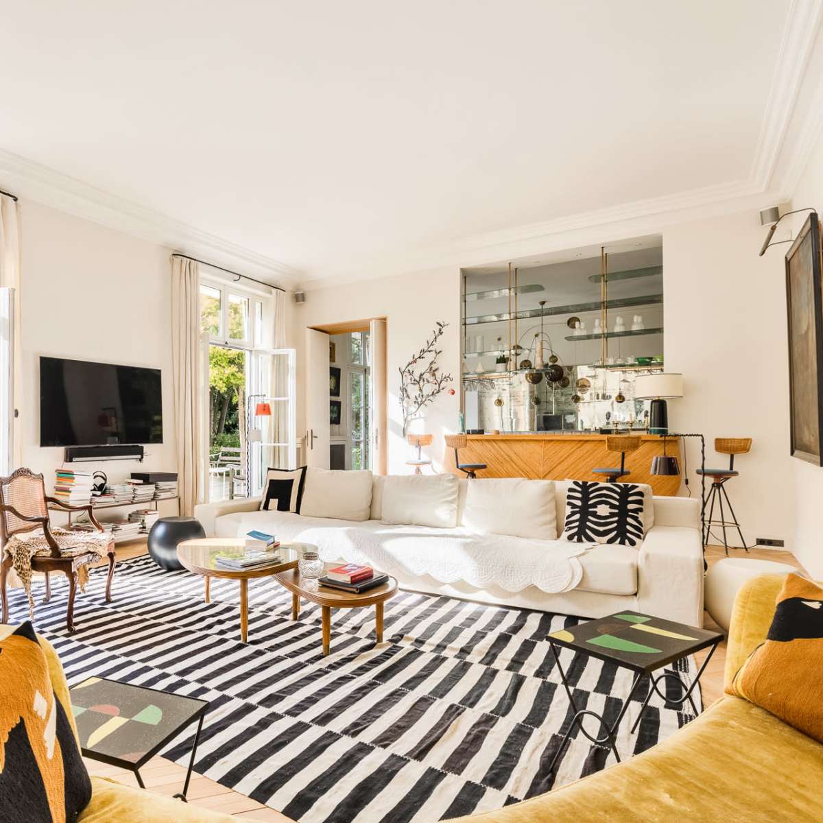 A bright living room with large windows, a comfortable sofa, a striped rug, and a wooden sideboard displaying decorative items.