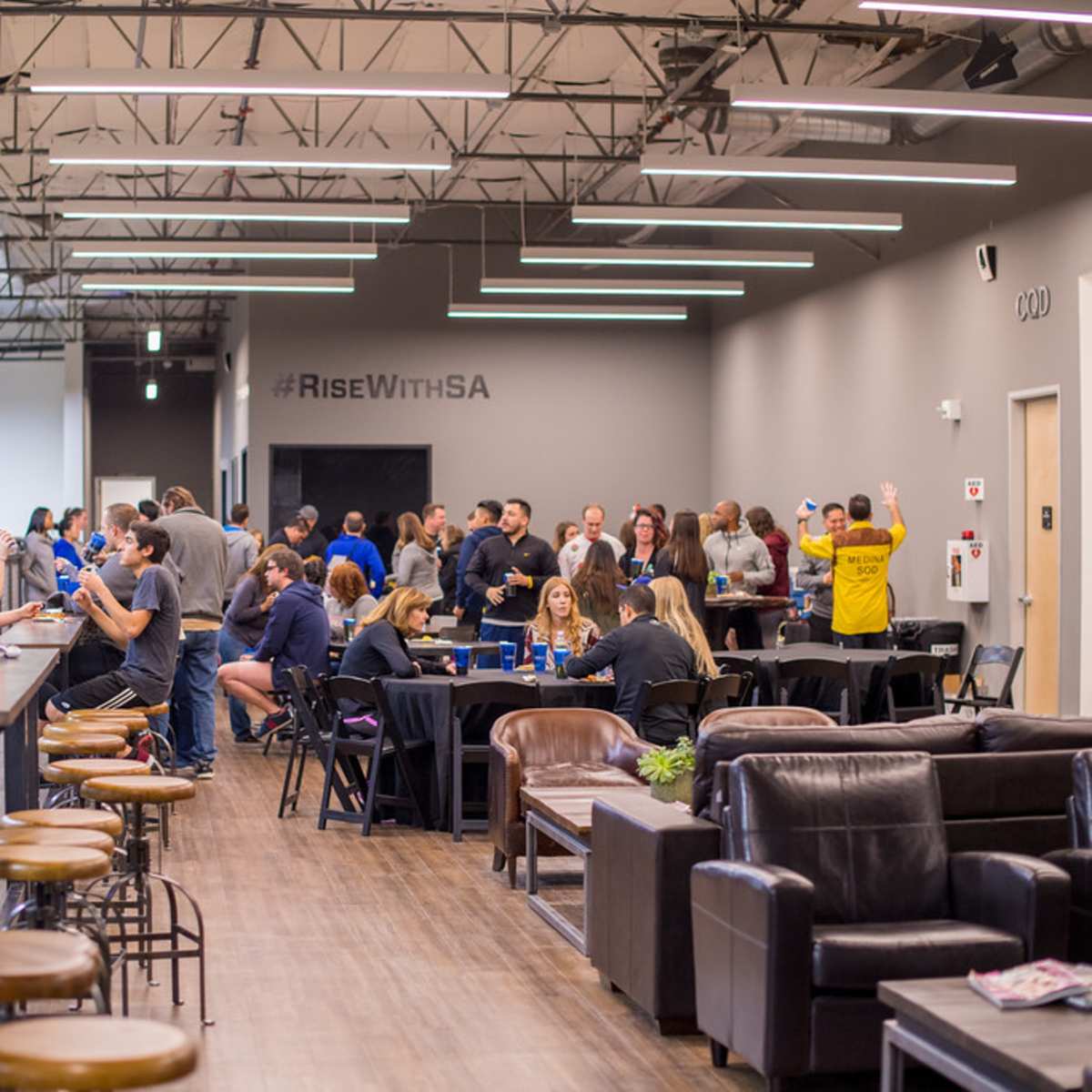 A group of people socializes in a spacious indoor venue with tables and seating, while a large wall displays the hashtag "#RiseWithSA."