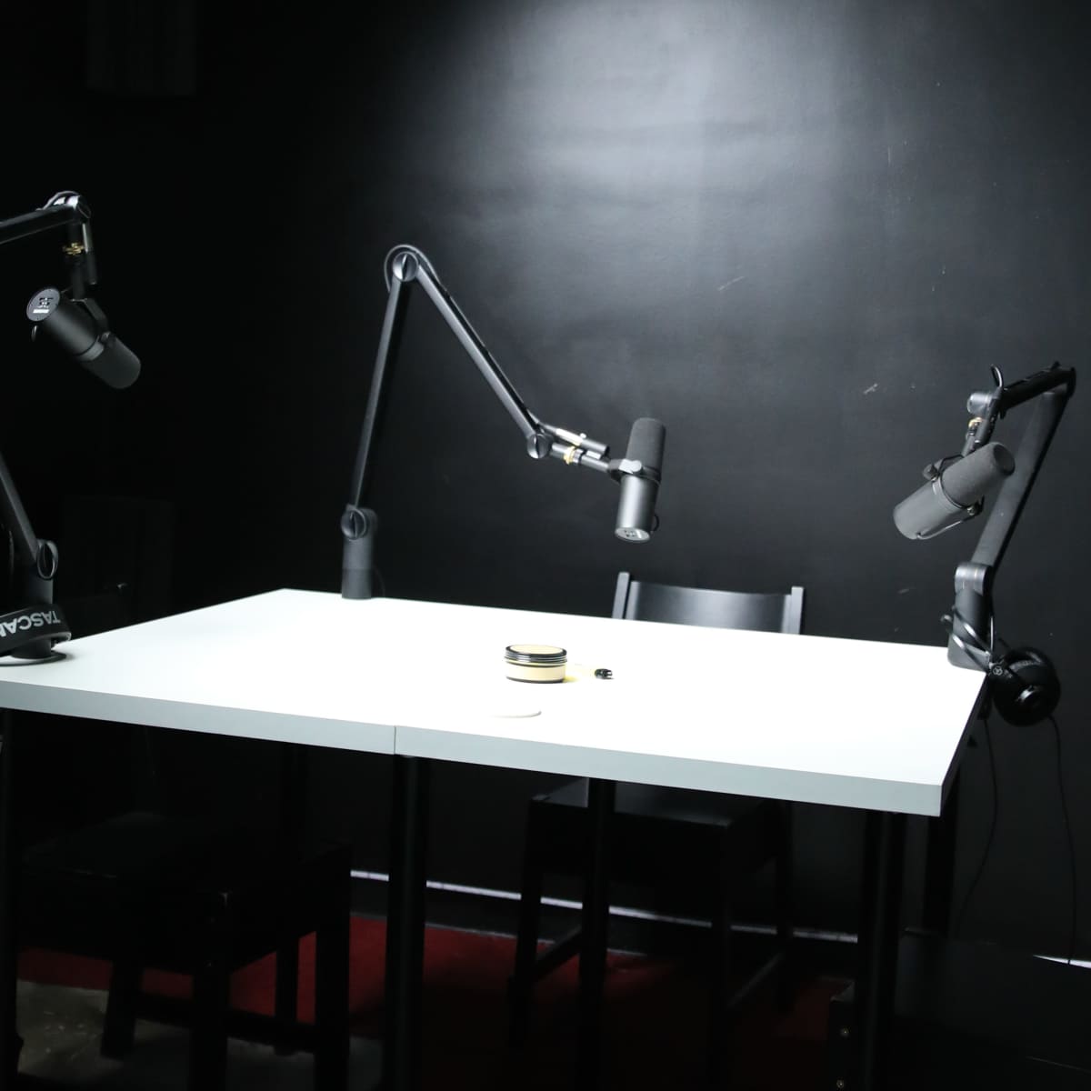 A minimalist recording studio featuring a white table with two adjustable microphones and two black chairs positioned around it, against a dark backdrop.