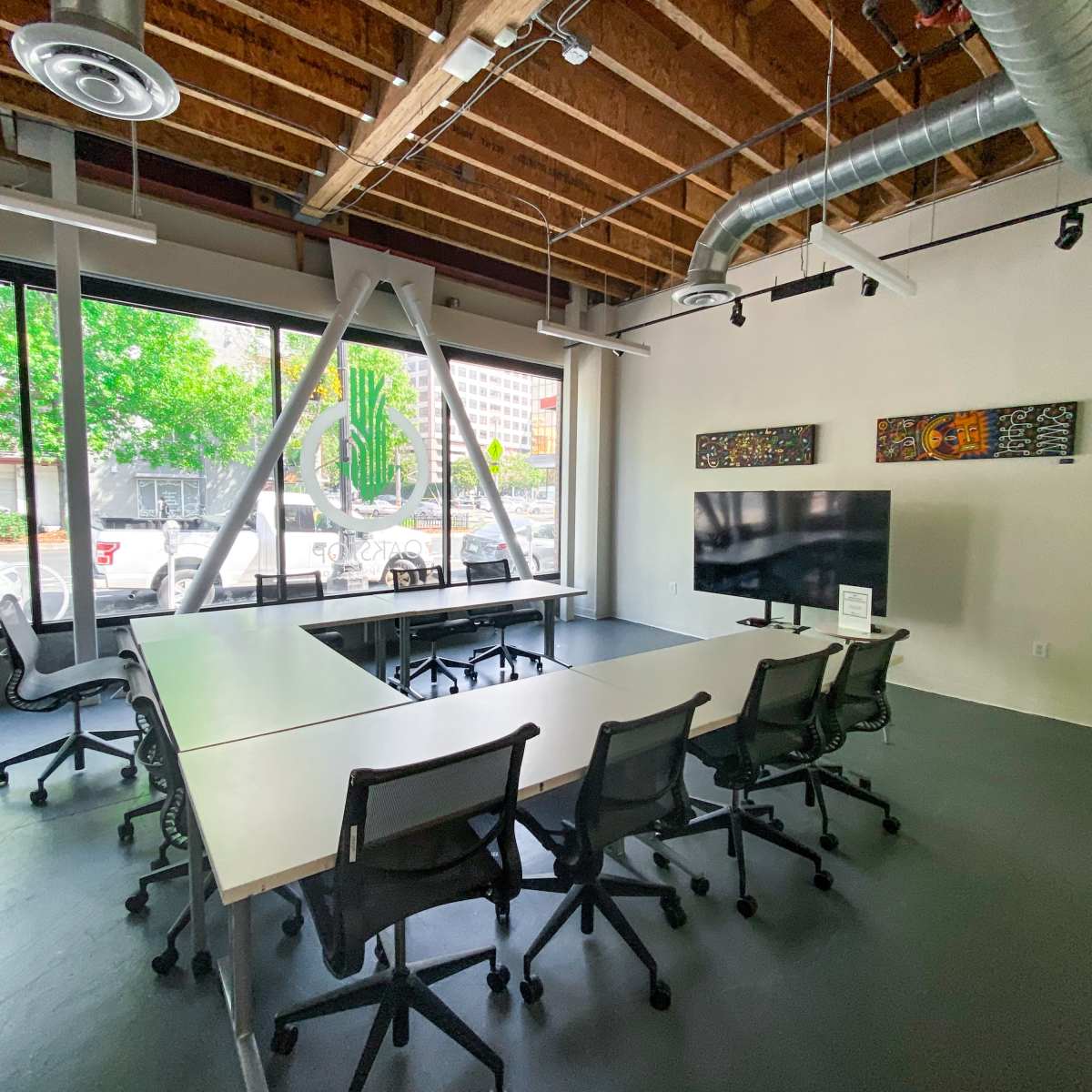 A modern conference room with a large table, several chairs, and a large screen, featuring exposed wooden beams and large windows overlooking a street.