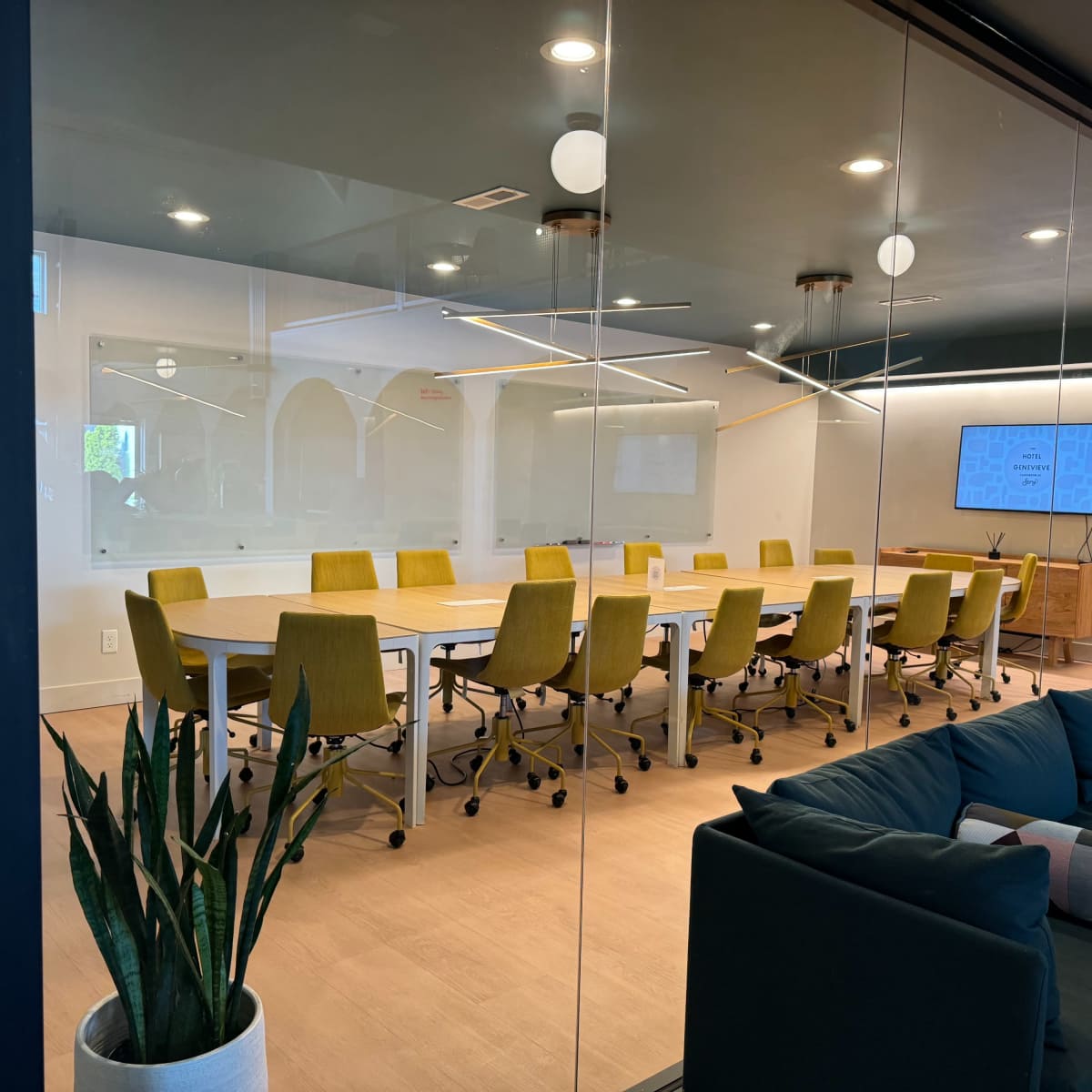A modern conference room features a long table surrounded by yellow swivel chairs, with a large screen on one wall and plants adding greenery to the space.