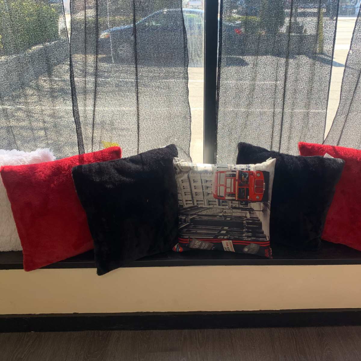 A row of decorative pillows in red, black, and white is arranged on a windowsill with sheer curtains behind them.