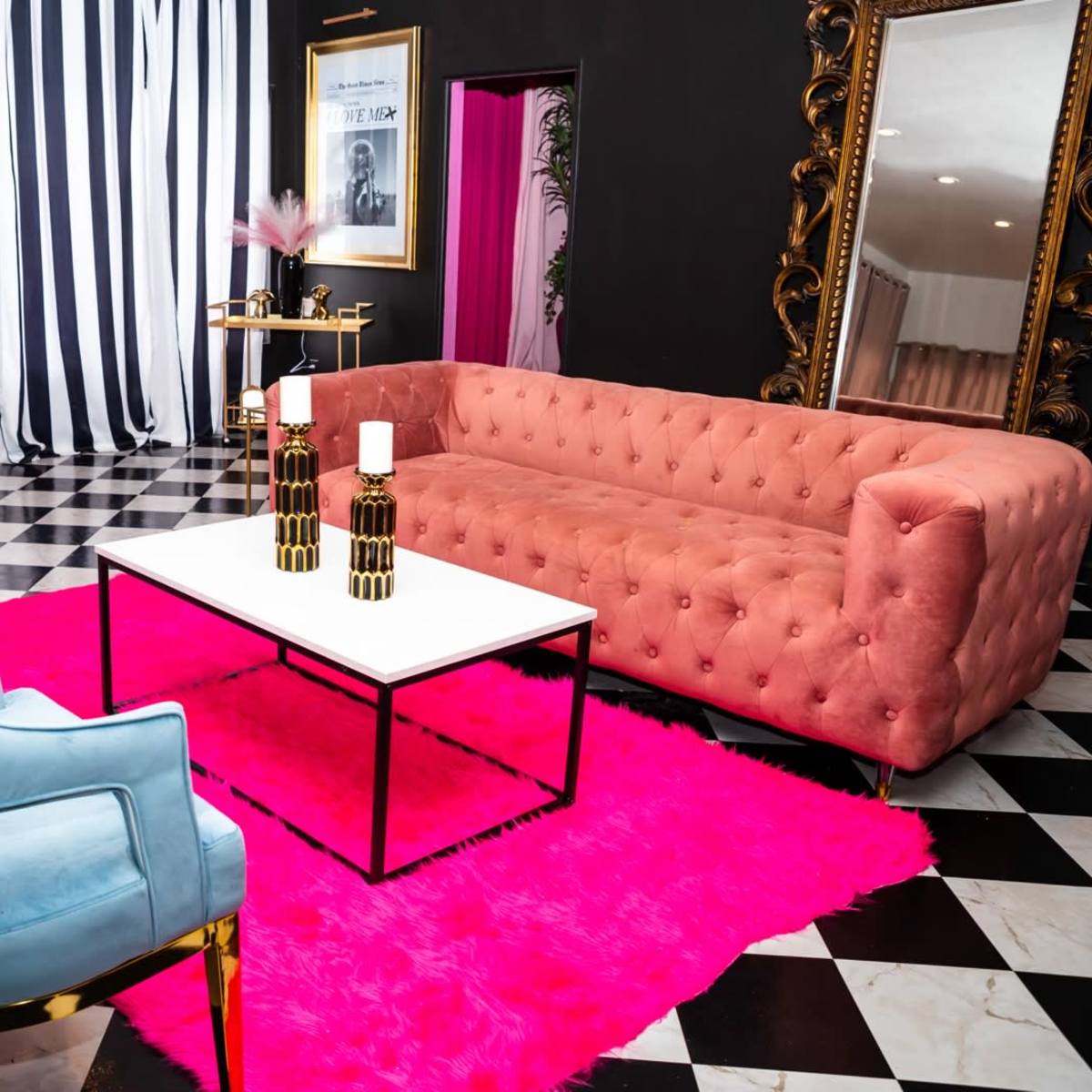 A living room features a pink sofa, blue chairs, a black and white checkered floor, and a white coffee table on a bright pink rug.