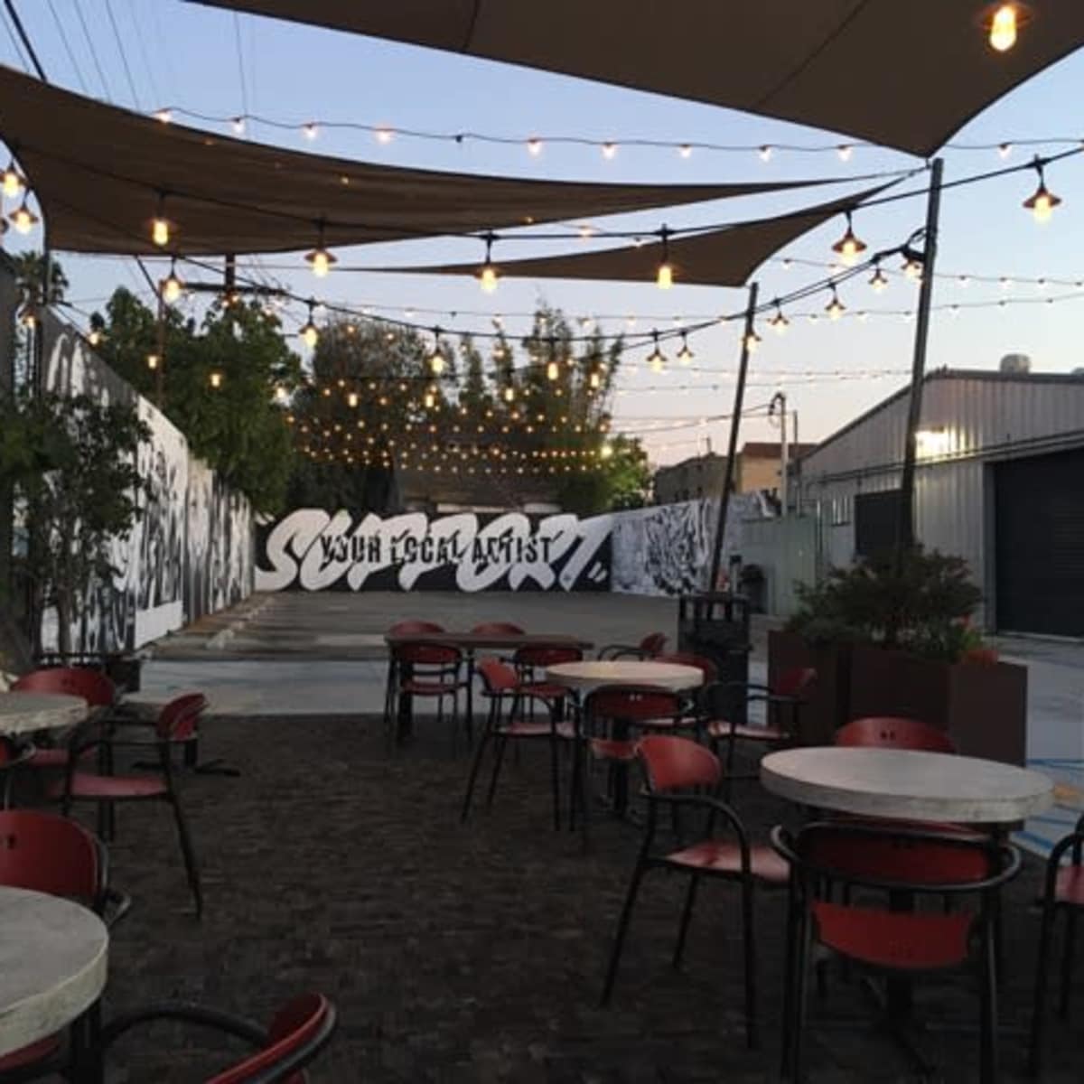 An outdoor seating area with tables and chairs beneath string lights, adjacent to a wall featuring street art.