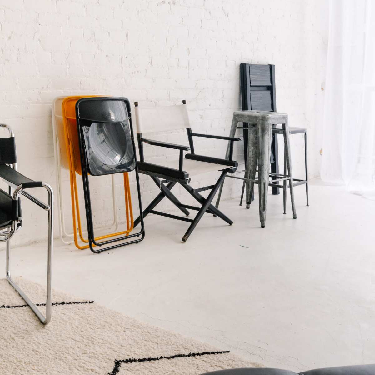 The scene features a variety of chairs, including stacked folding chairs, a bar stool, and a director's chair, against a white brick wall.