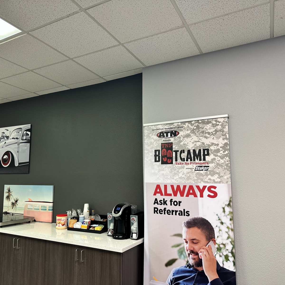 An office space featuring a dark gray wall with a framed car photo, a countertop displaying coffee equipment and snacks, and a banner promoting referrals.