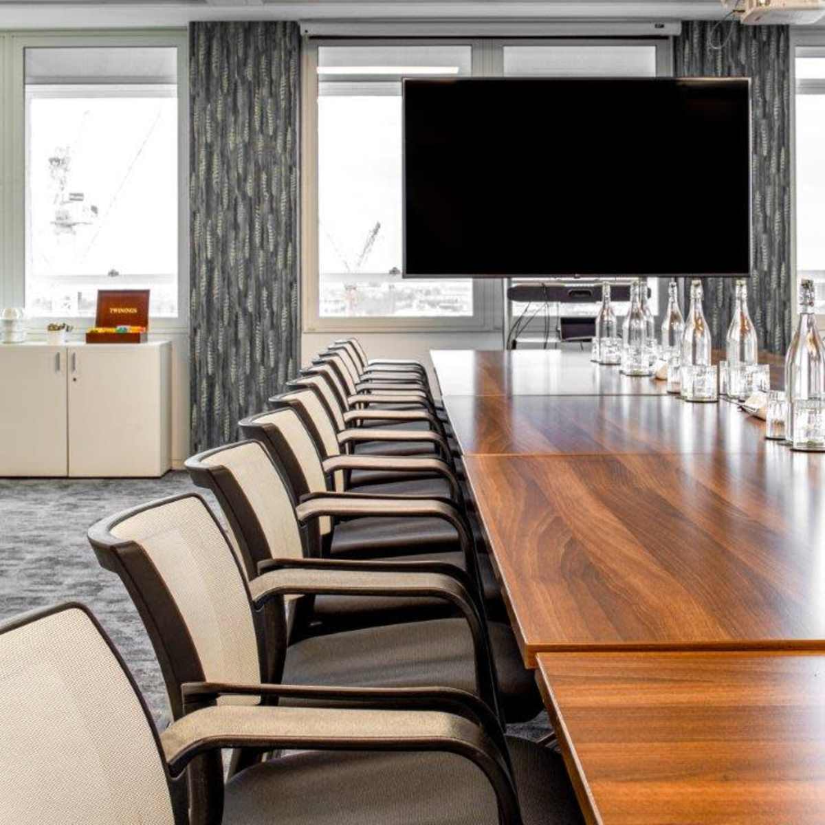 A long conference table with several chairs is set up in a modern meeting room featuring large windows and a wall-mounted screen.