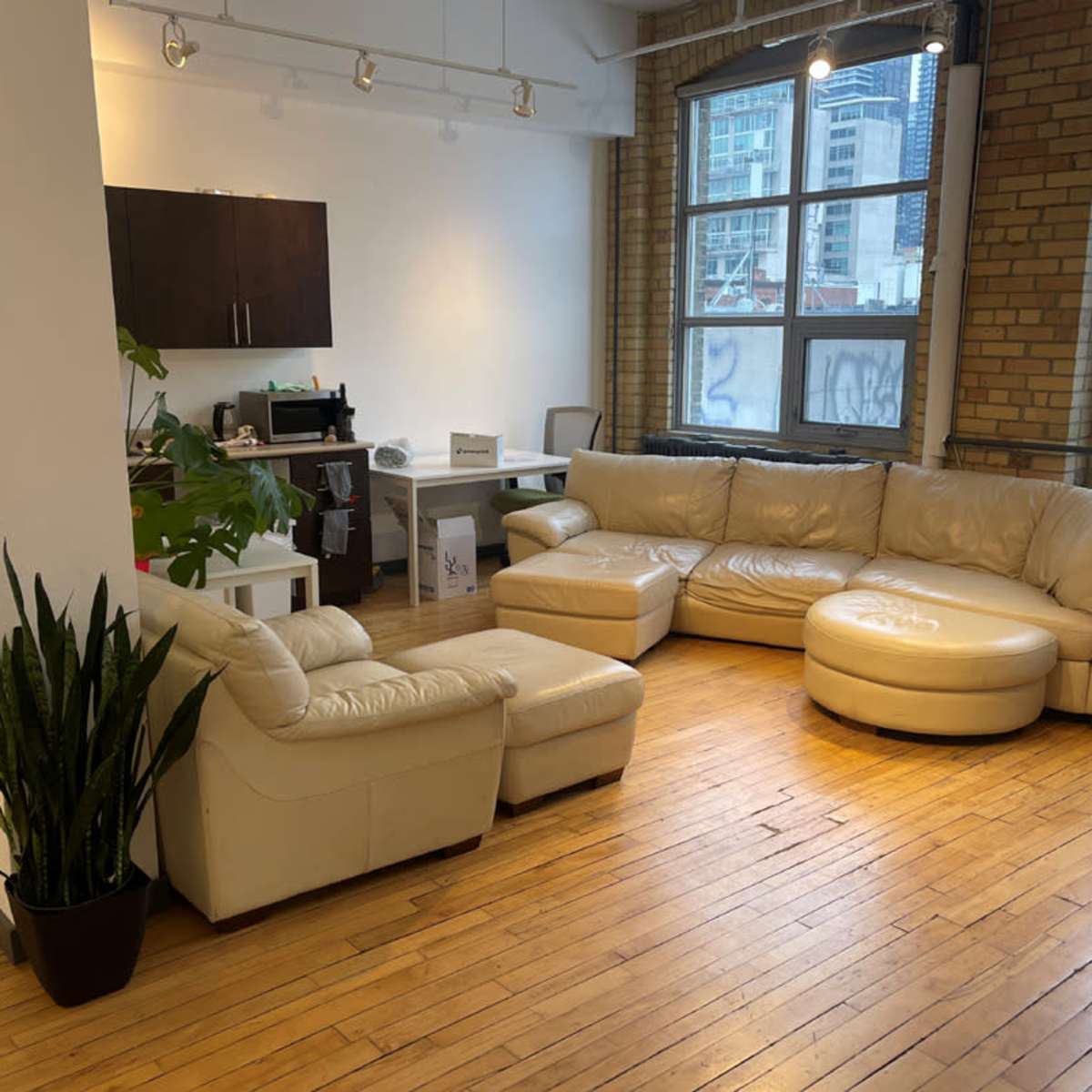 A spacious room with a large beige leather sectional sofa, a white desk, and large windows overlooking a cityscape.