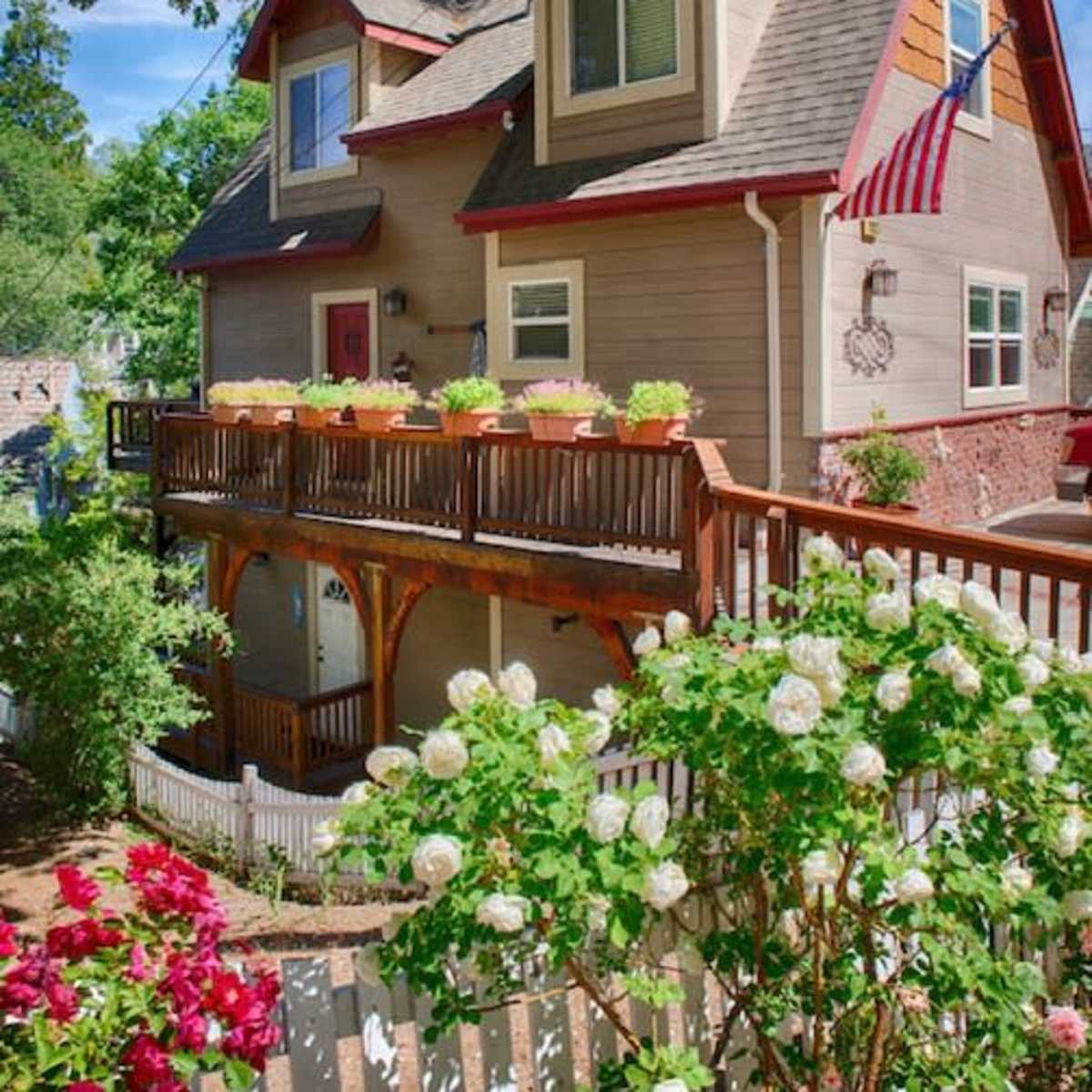 A two-story house with a wooden deck adorned with flower pots and a flag, surrounded by a garden featuring rose bushes and a white picket fence.