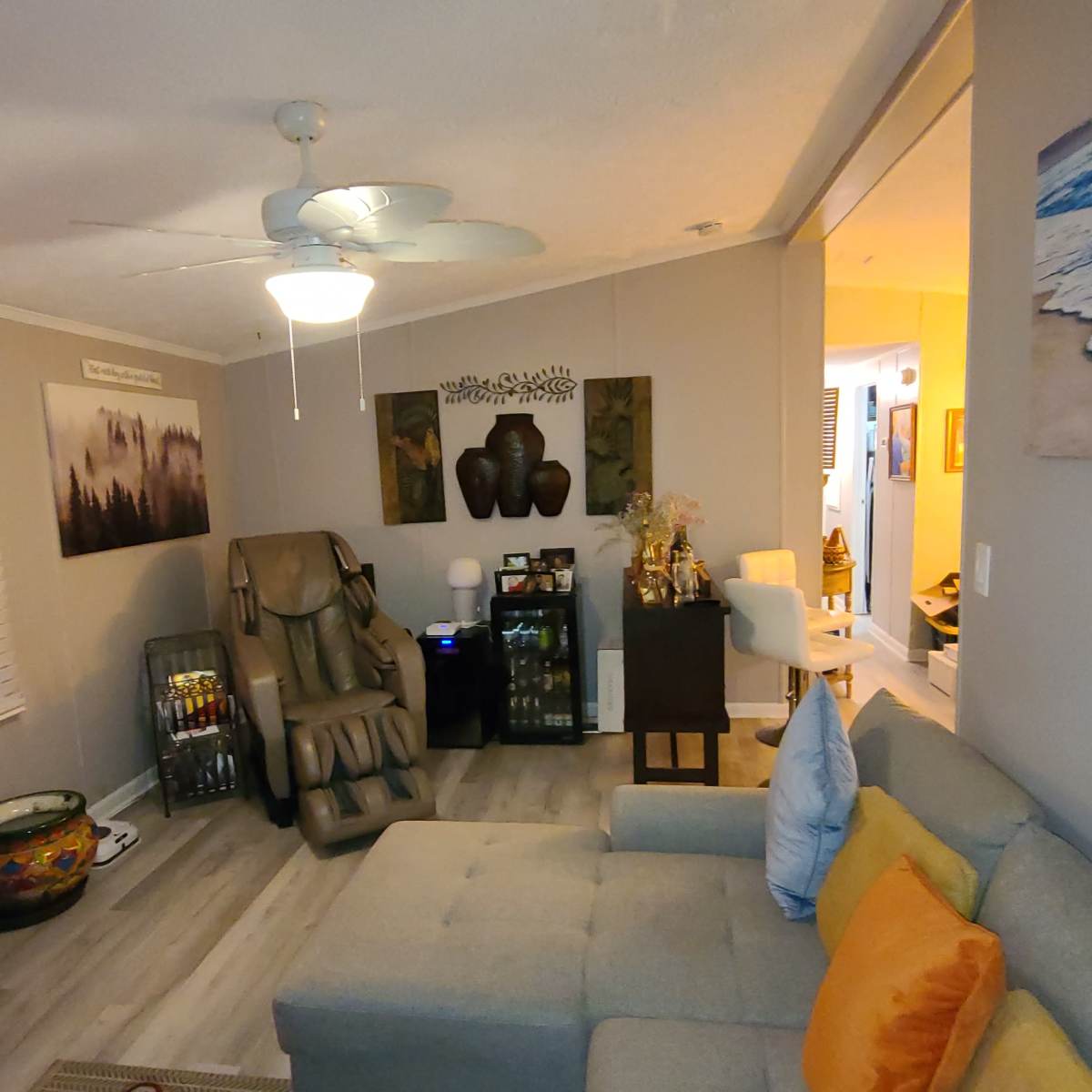A cozy living room with a couch, a massage chair, and a small table, illuminated by a ceiling fan and soft lamps.