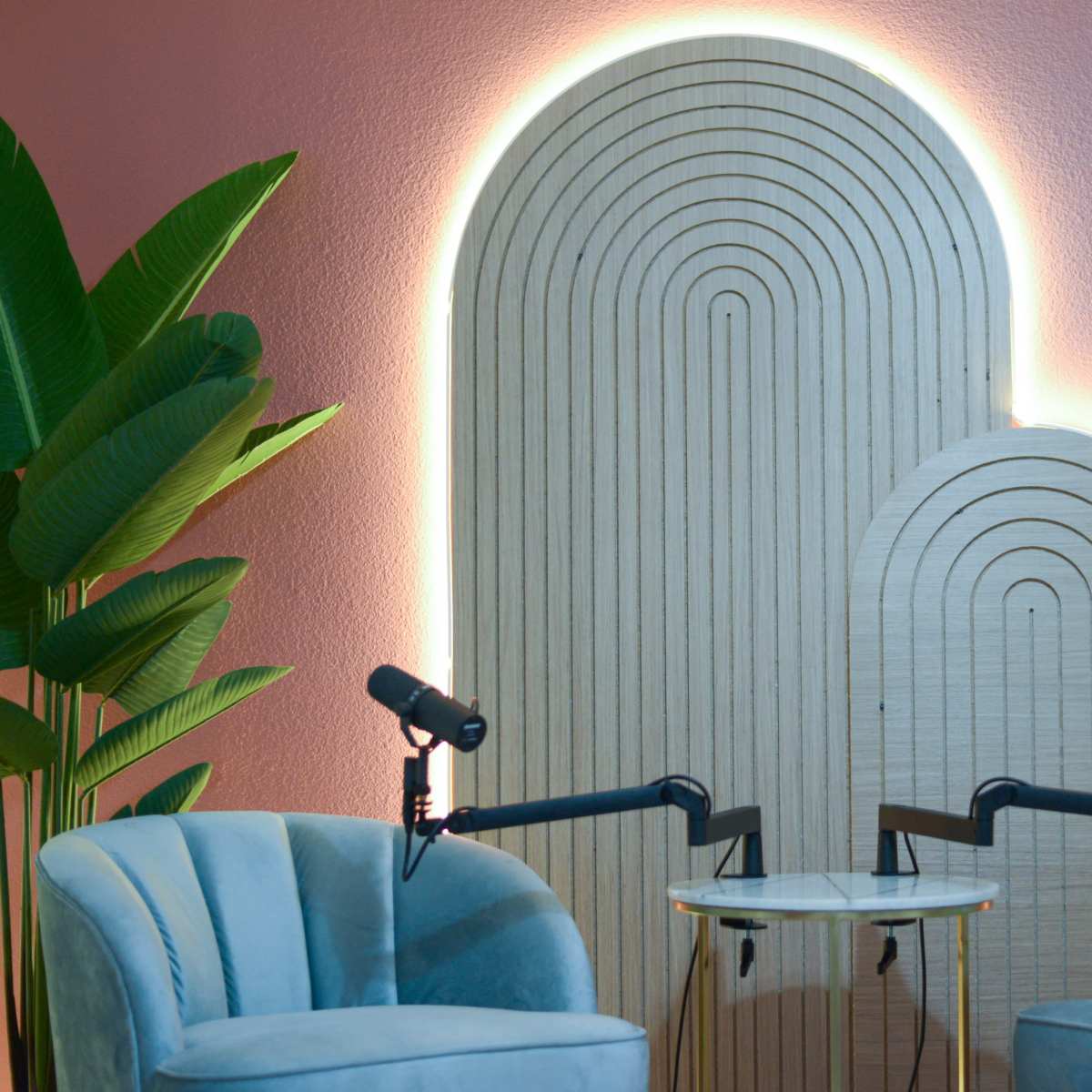 Two blue chairs facing each other with a small table between them, set against a backdrop of layered wooden arches illuminated by soft lighting, surrounded by green plants.