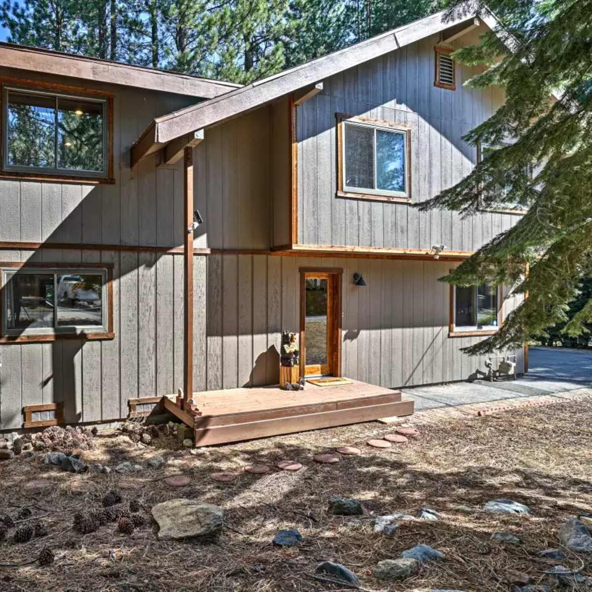 A two-story wooden house with a light gray exterior, surrounded by pine trees and a gravel pathway.