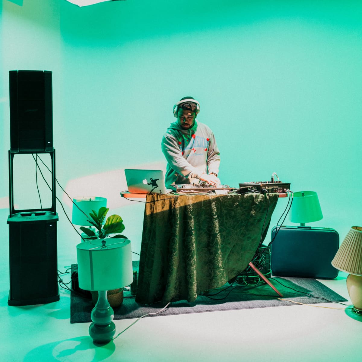 A DJ stands behind a table covered with a cloth, mixing music in a studio with green lighting and various decorative items around him.