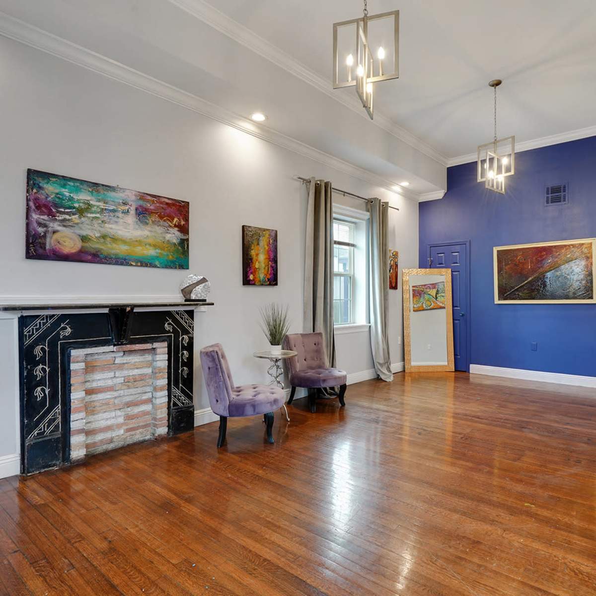 A spacious living room featuring wooden floors, a decorative fireplace, vibrant artwork on the walls, and large windows allowing light to enter the area.