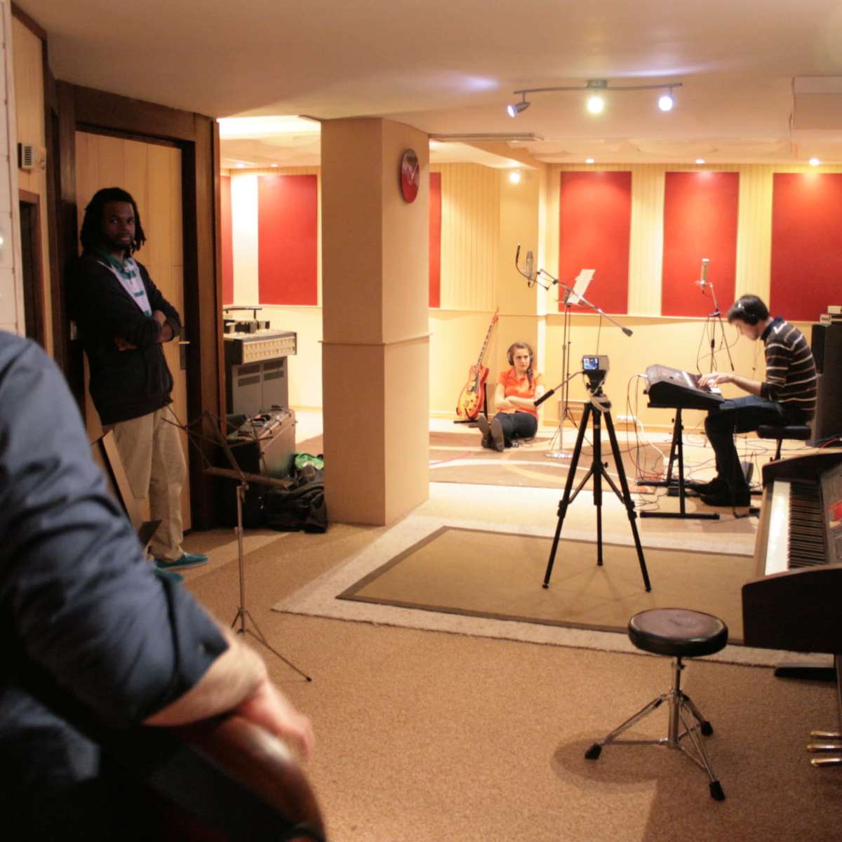 Musicians are seen rehearsing in a soundproof studio, with one playing guitar and another seated at a piano, while a microphone and cameras are set up for recording.