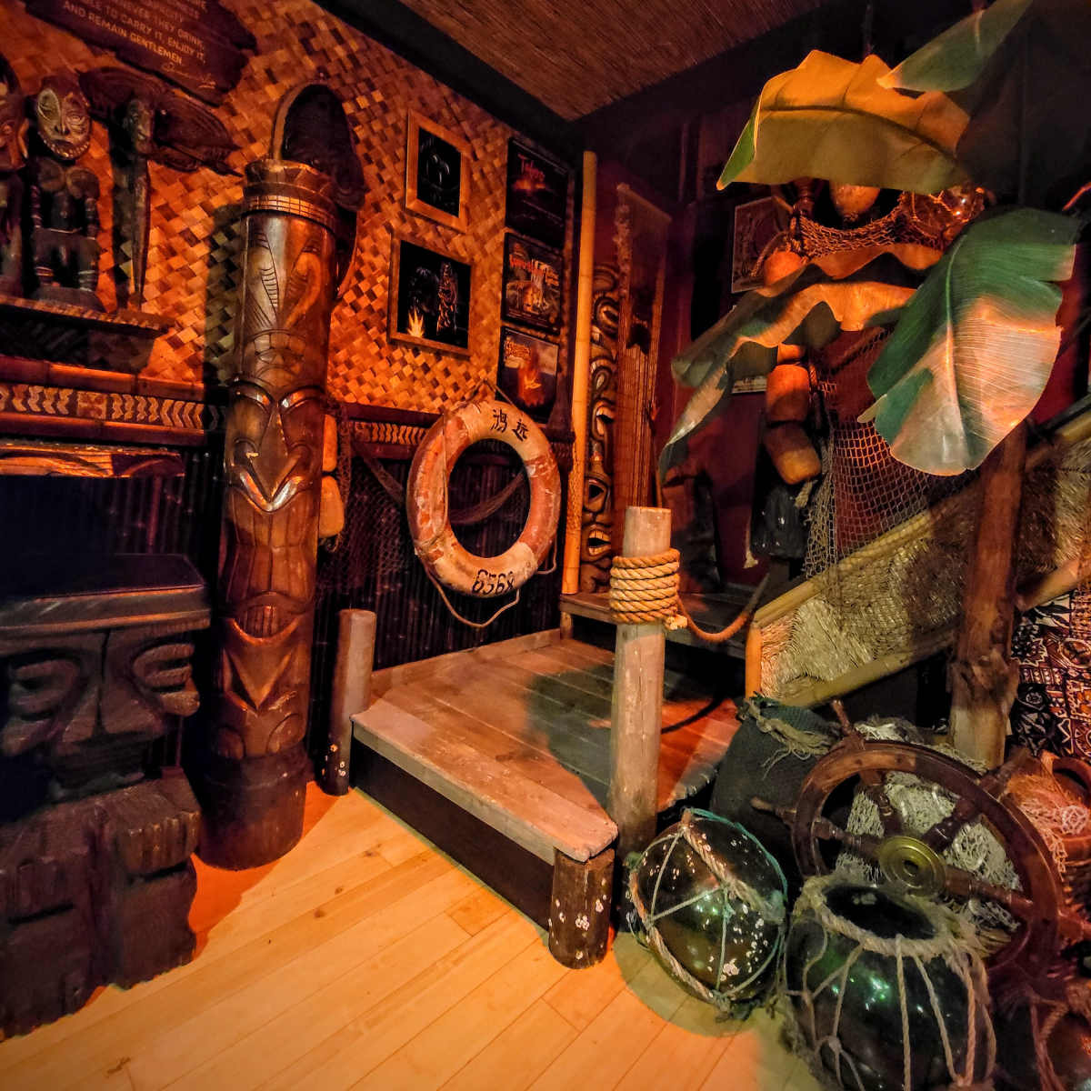 A dimly lit room featuring a wooden platform surrounded by tropical decorations, carved wooden figures, and various artifacts.