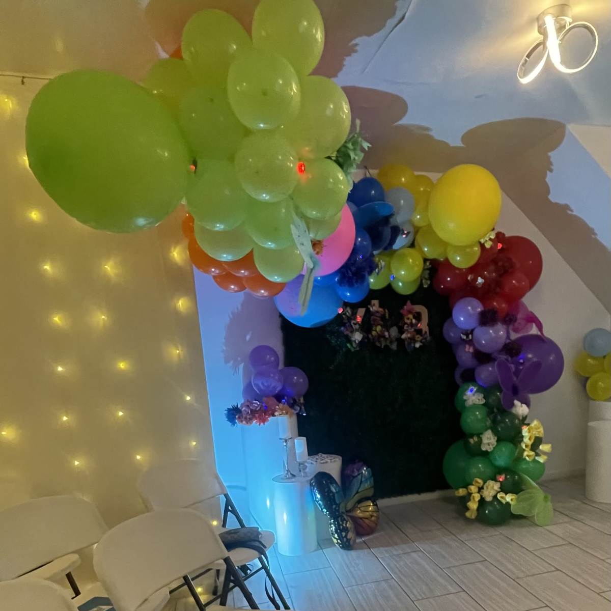 A cluster of colorful balloons is arranged in various shades near a wall adorned with string lights and a green backdrop.