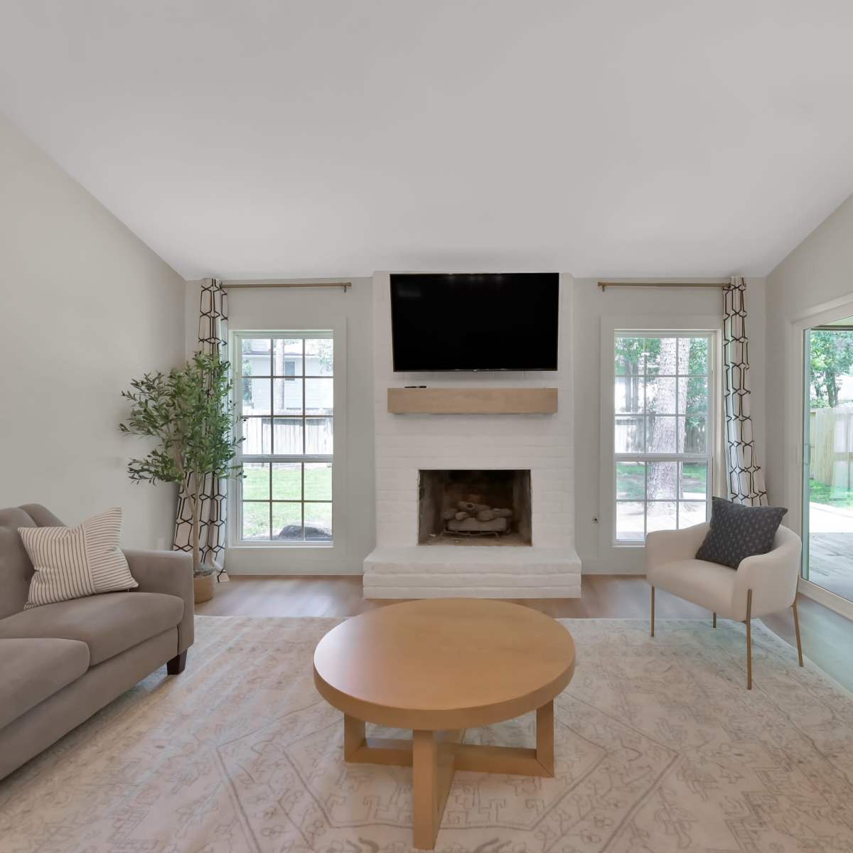 A living room with a gray sofa, a round wooden coffee table, a fireplace underneath a mounted TV, and large windows overlooking a backyard.