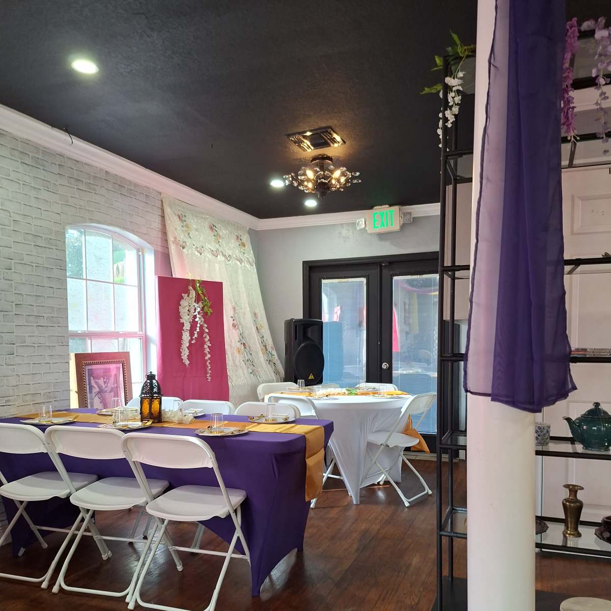 An elegantly decorated event space featuring tables set with purple and white tablecloths, a speaker system, and decorative elements against a brick wall.