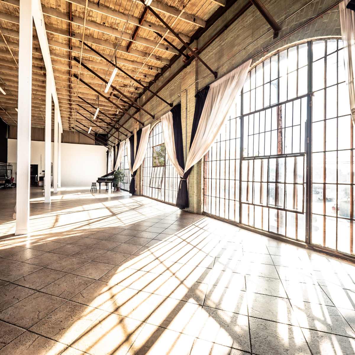 A spacious industrial room with large windows, wooden beams, and long shadows cast across the tiled floor.