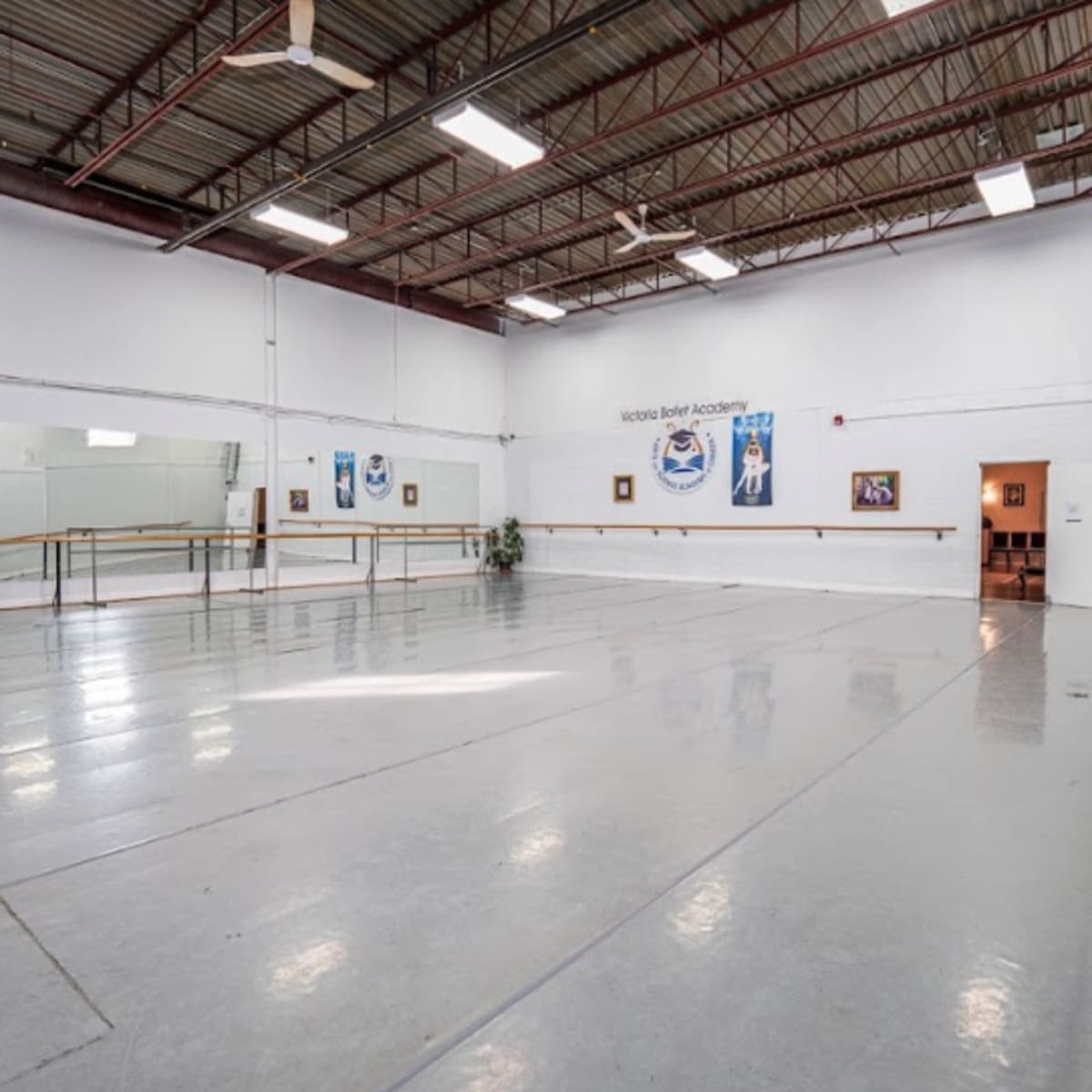 An empty dance studio features a mirrored wall, ballet barres, and a concrete floor, with artwork displayed on the walls.