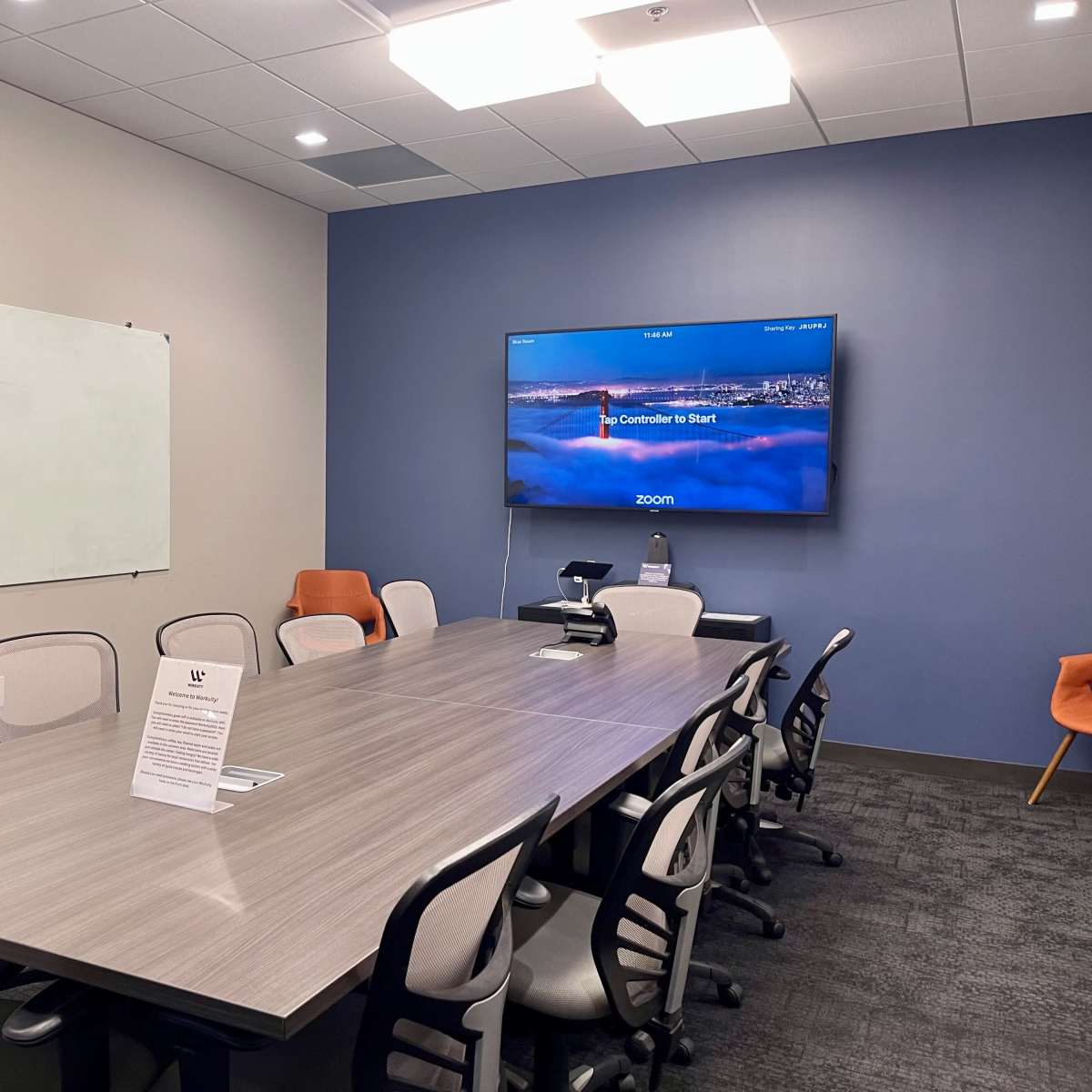 A conference room features a long, rectangular table surrounded by chairs, with a whiteboard on one wall and a screen displaying a Zoom interface on another.